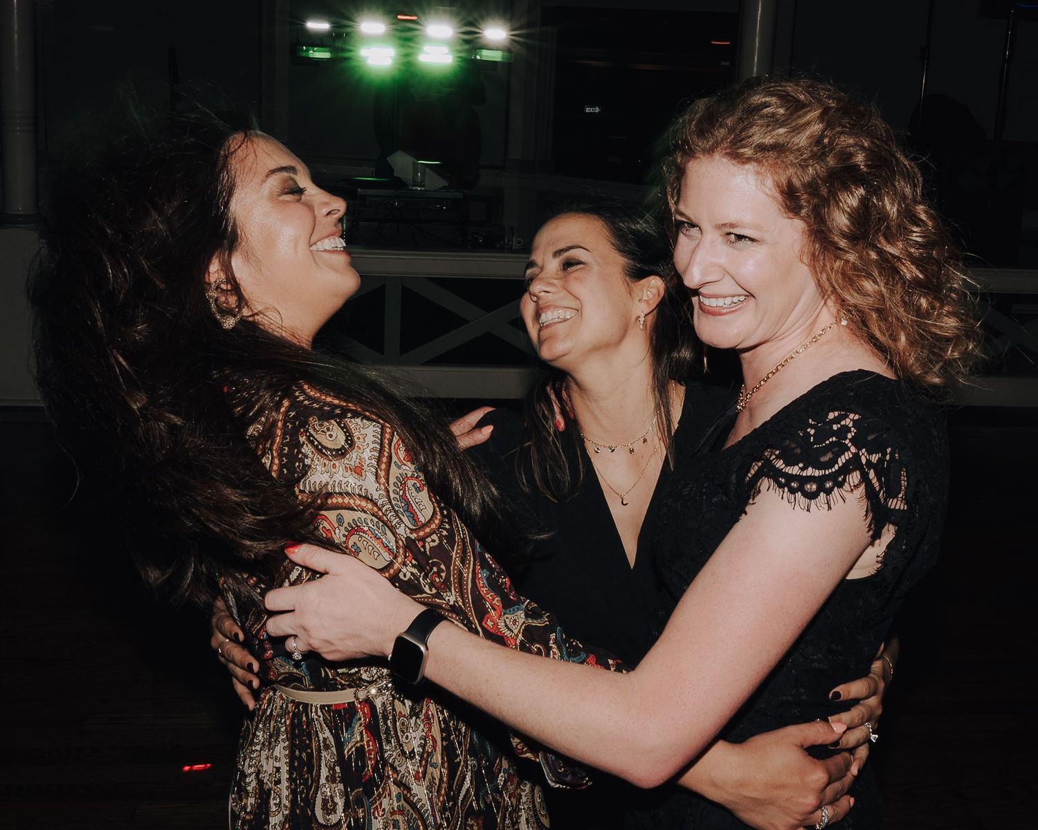 Three women laughing and embracing at an evening celebration event.