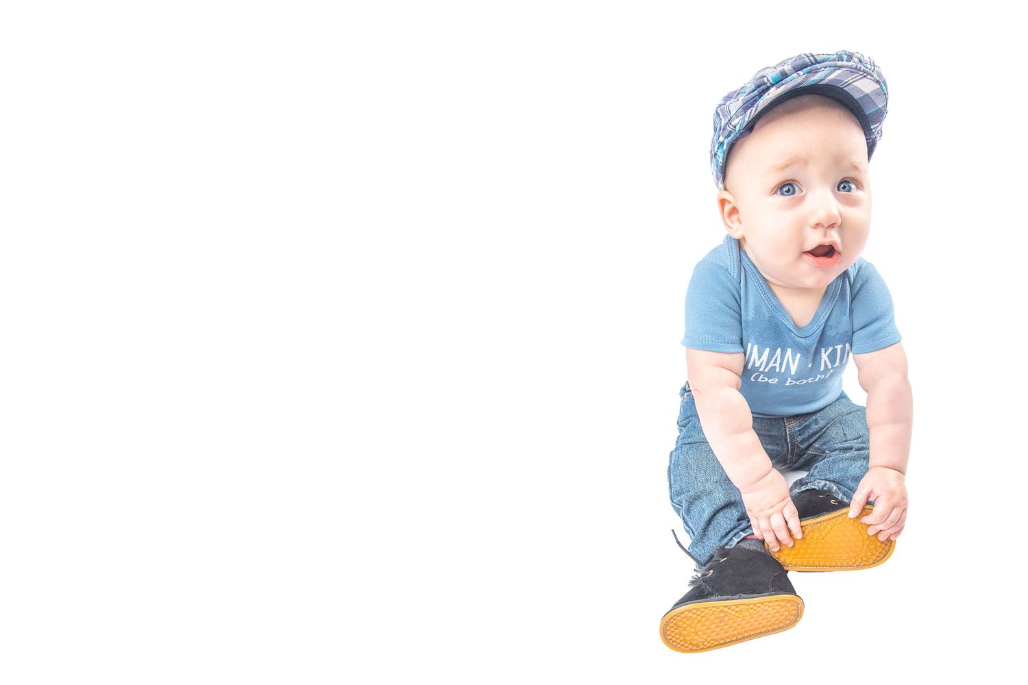 Cute baby boy in blue outfit and cap sitting on white background looking up curiously.