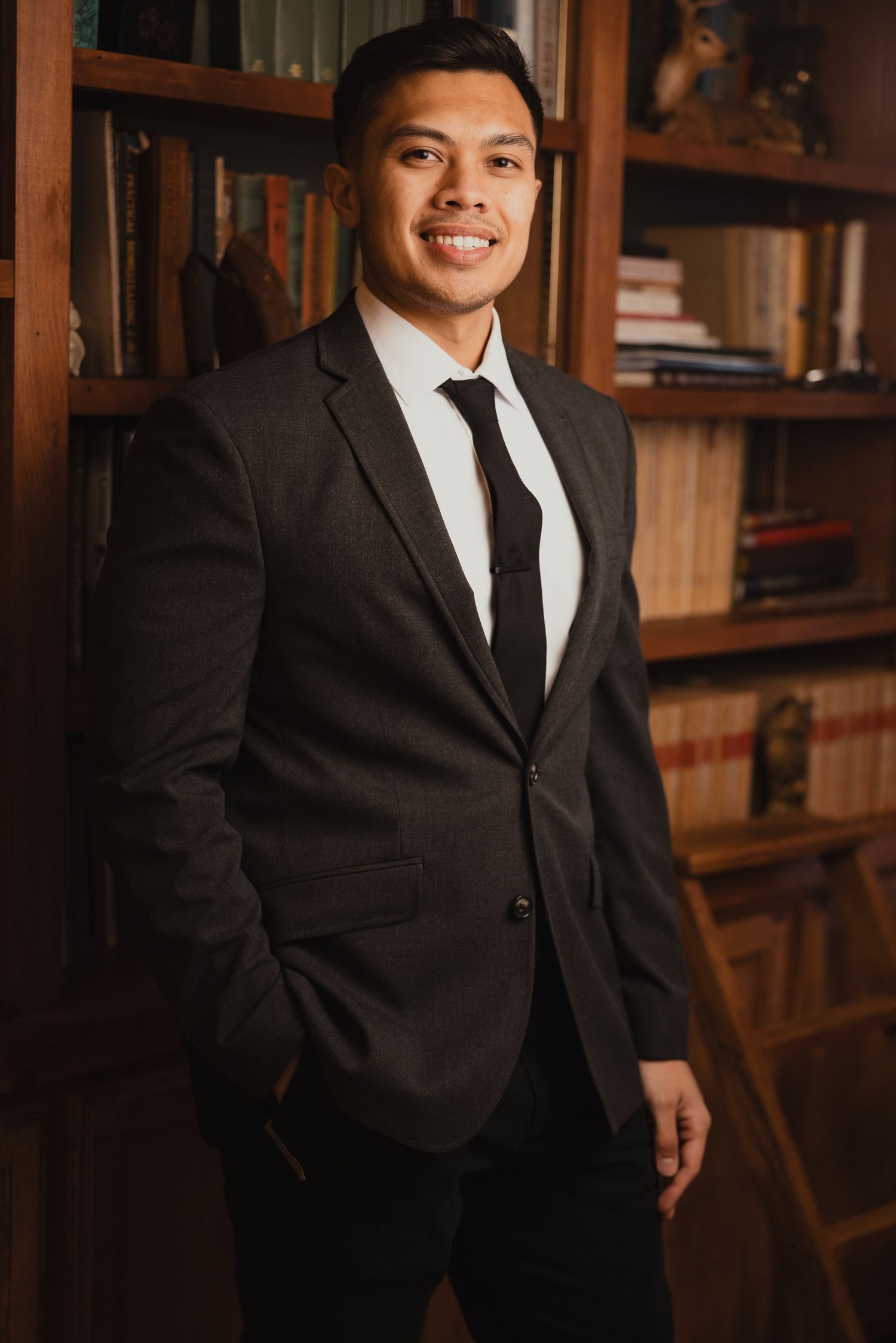 Professional portrait of Carl Michael in a black suit and tie standing in front of a wooden bookshelf in a library setting.