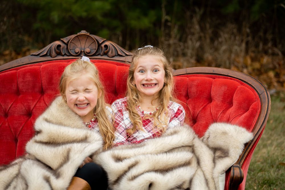 Two children wrapped in blankets laugh together on a vintage red velvet sofa outdoors.