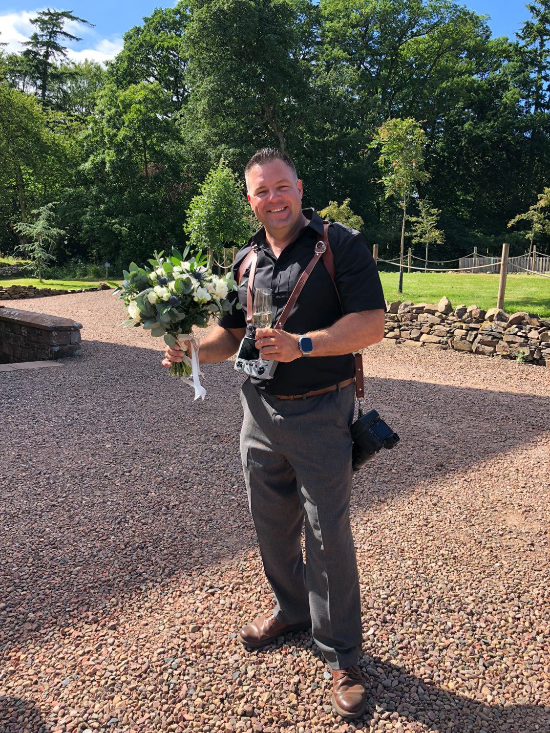 Professional photographer in black attire holding camera equipment and bouquet outdoors.