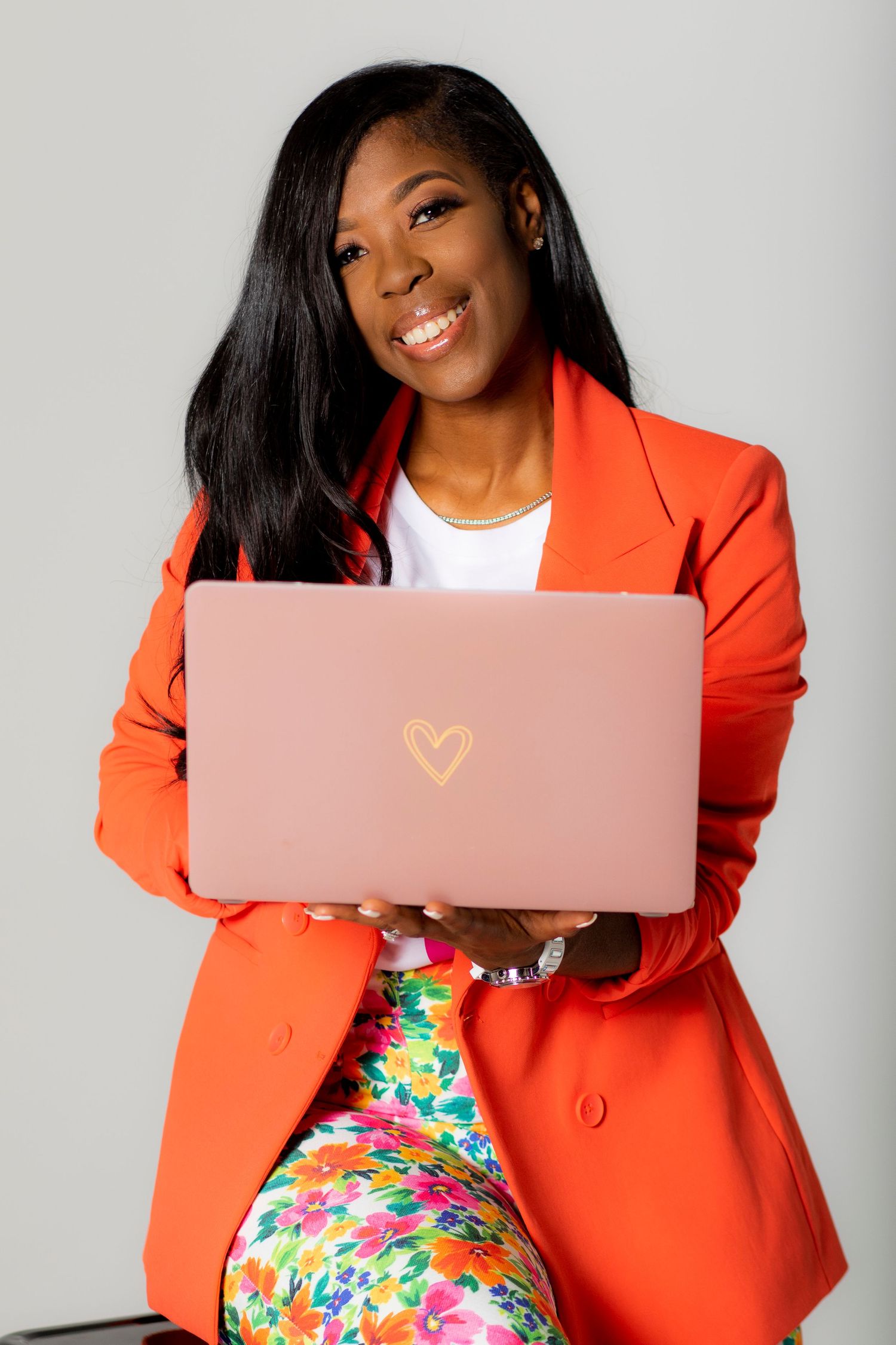 Professional entrepreneur smiles while holding laptop with heart detail in bright orange blazer.