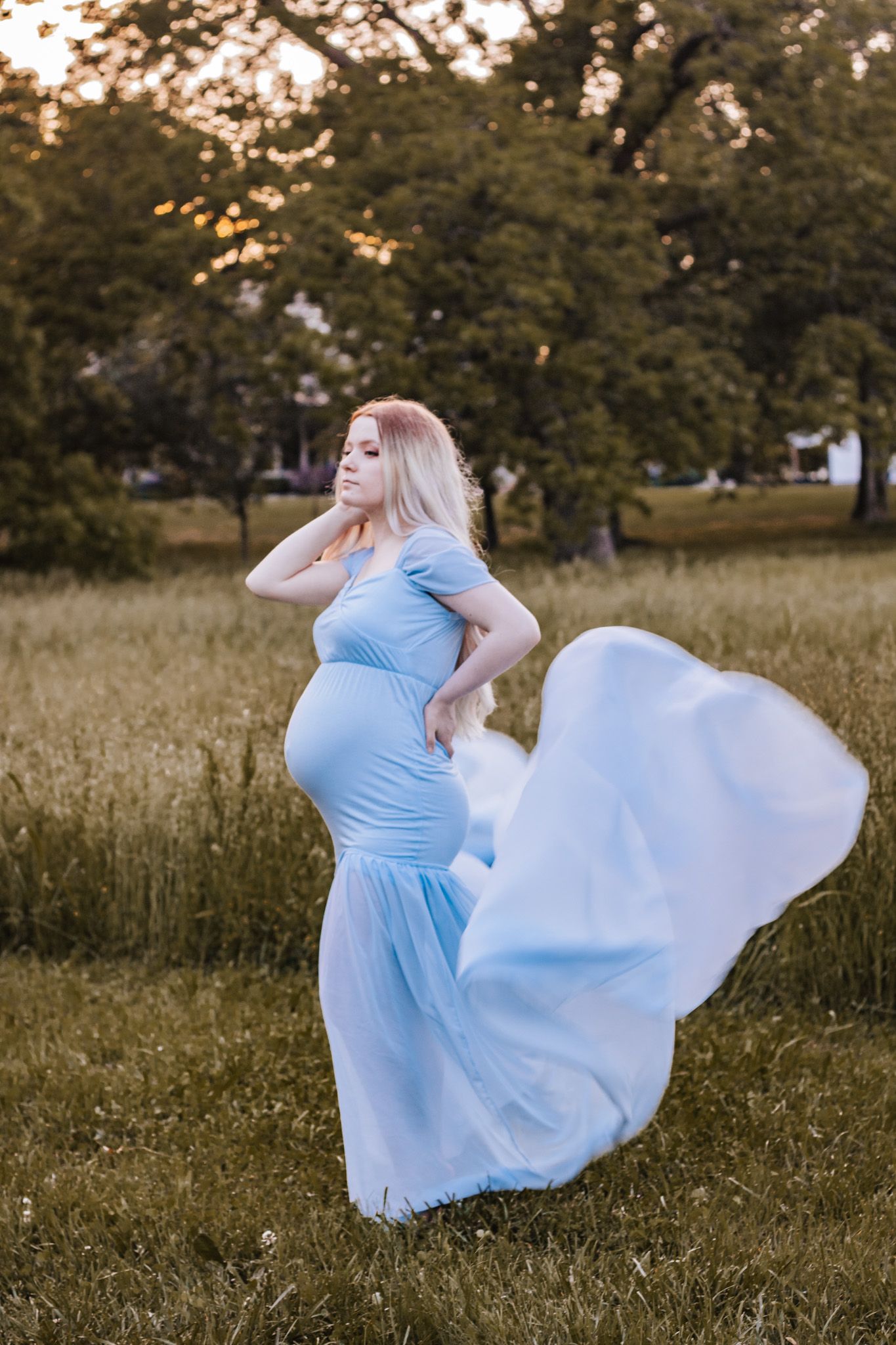 Woman in flowing light blue dress twirling in sunlit meadow at sunset.