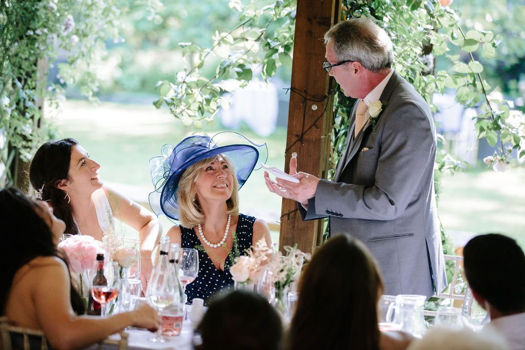 Father of the bride doing his speech with mother of the bride and bride smiling at him.