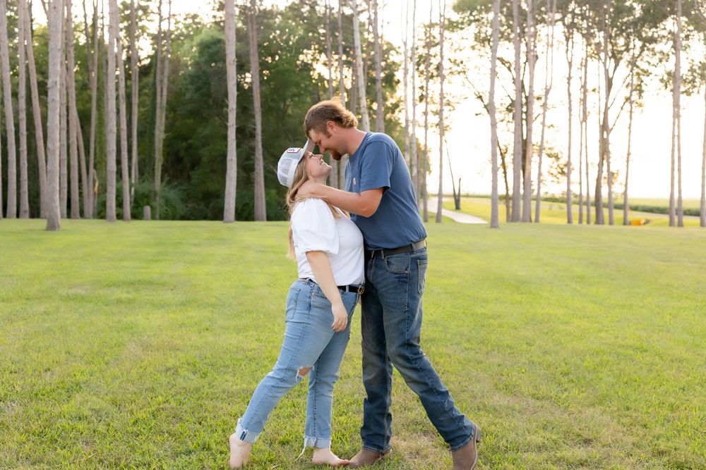 A couple embracing on a grassy field with pine trees silhouetted in the evening light.