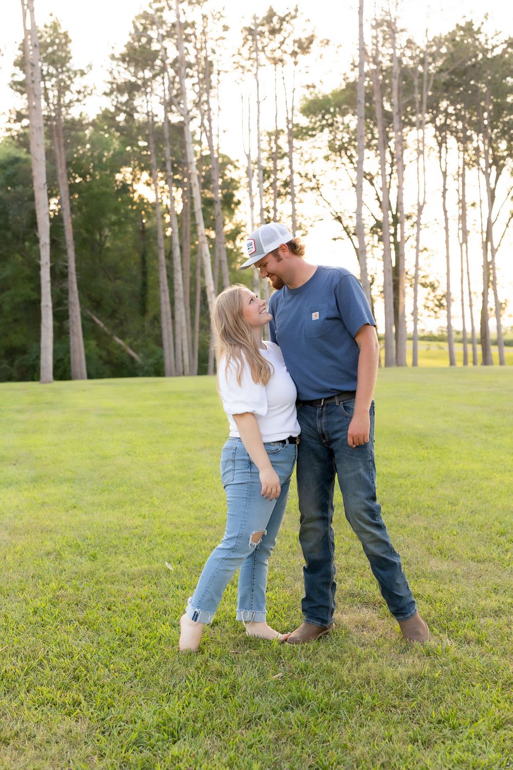 A couple stands close together on a grassy field with tall trees in the background during golden hour.