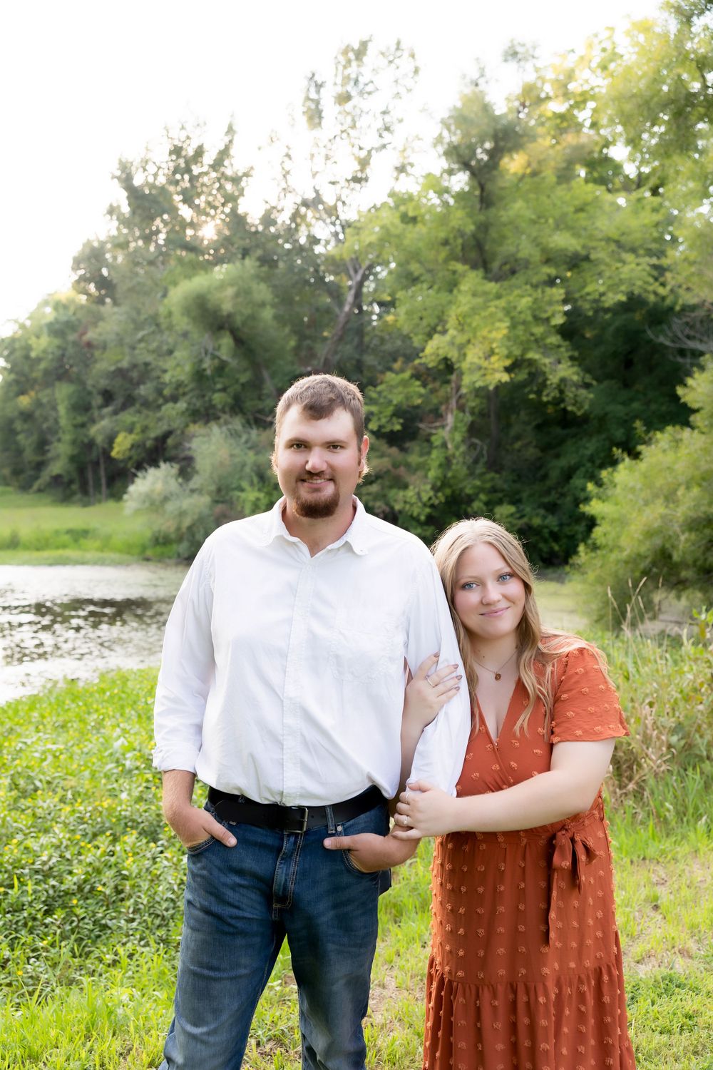 A couple poses together by a pond with lush greenery in the background on a sunny day.