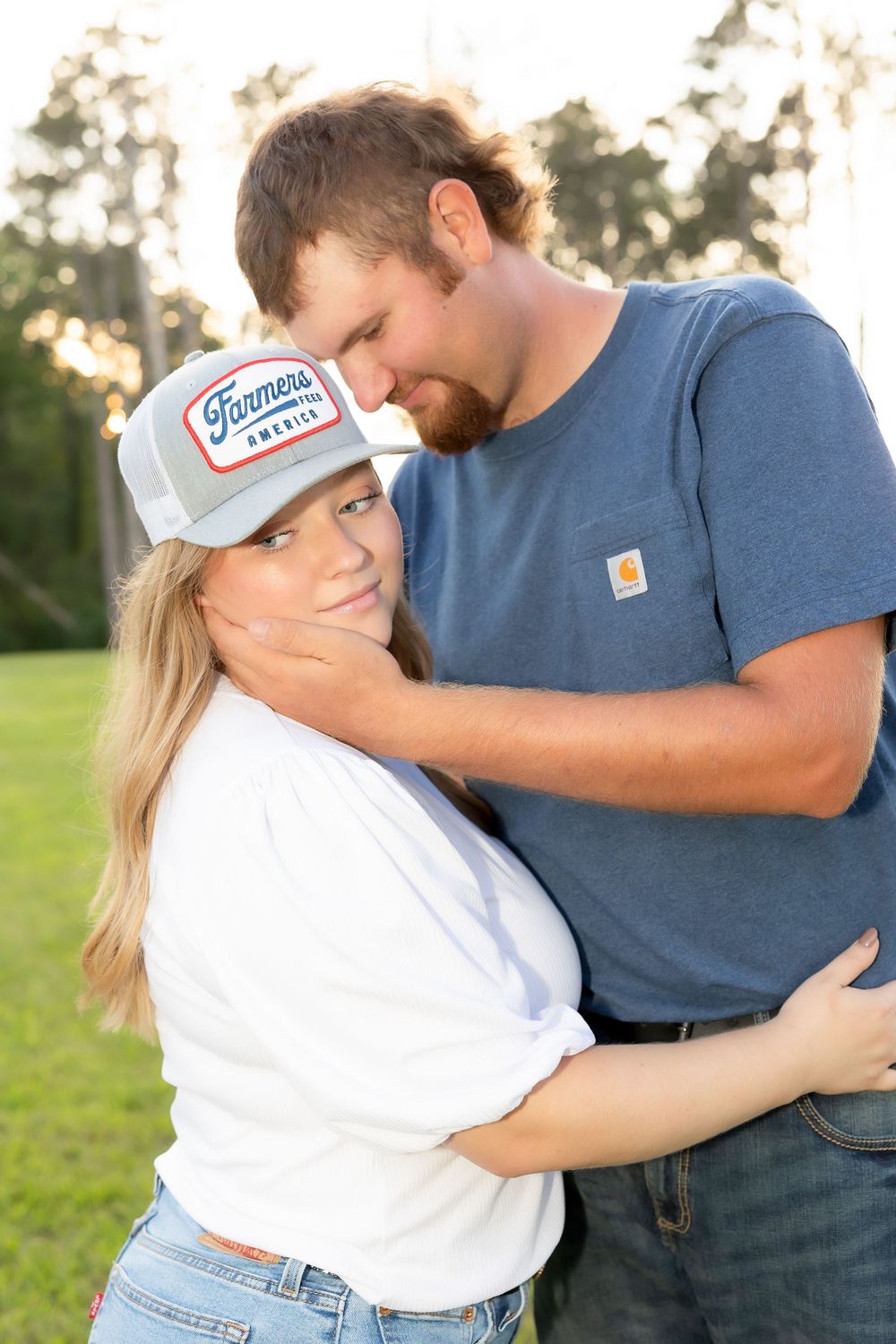 A couple shares an intimate moment outdoors, with one wearing a white baseball cap.