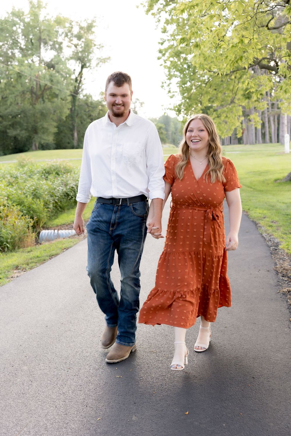 A couple strolling down a tree-lined path, with her wearing a rust-colored dress and him in jeans and white shirt.