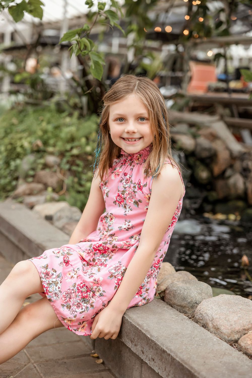 A young girl in pink floral dress sits on stone border in a garden setting.