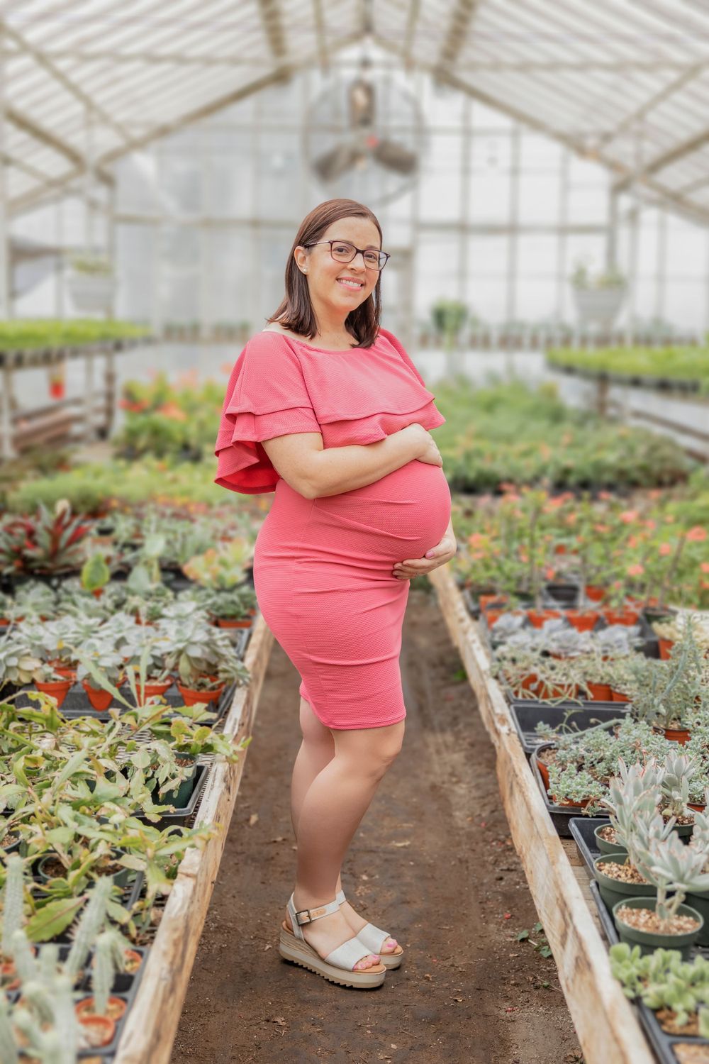 Expectant mother in coral dress poses in greenhouse walkway surrounded by plants.