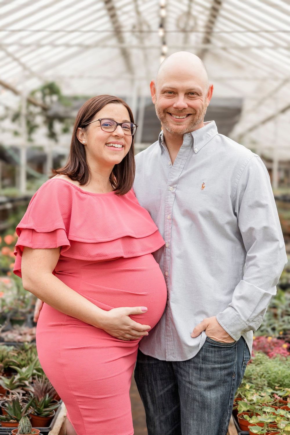 Expectant couple poses together in greenhouse surrounded by plants and flowers.