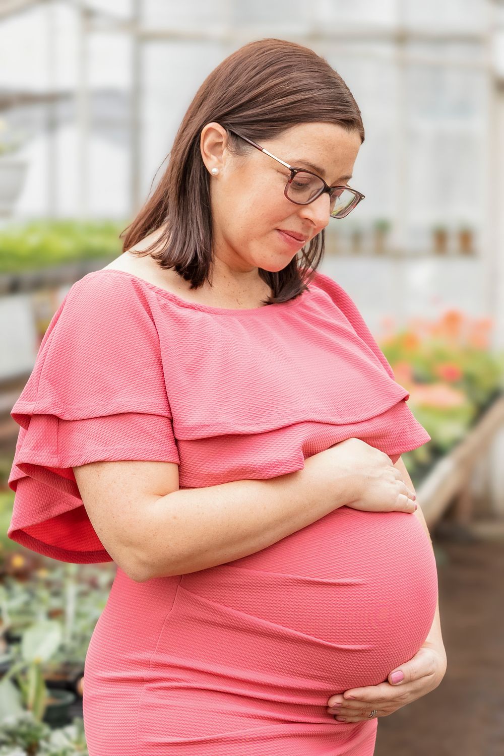 Expectant mother in coral ruffle dress poses while cradling her baby bump in greenhouse.