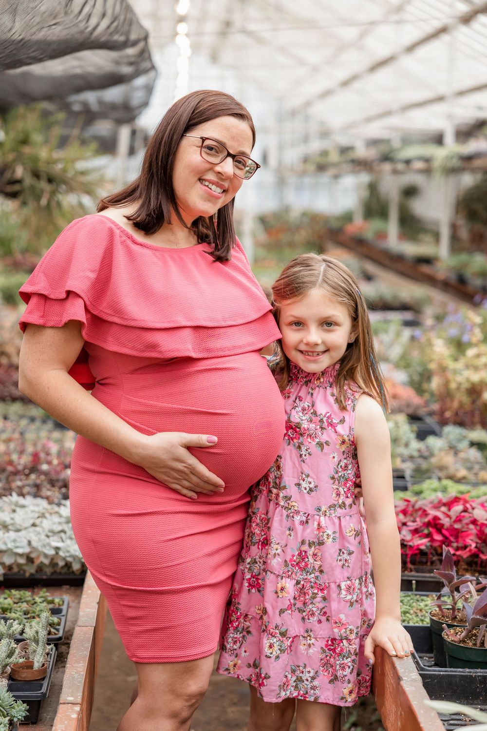 An expectant mother in coral dress and young girl in floral dress pose together in greenhouse.