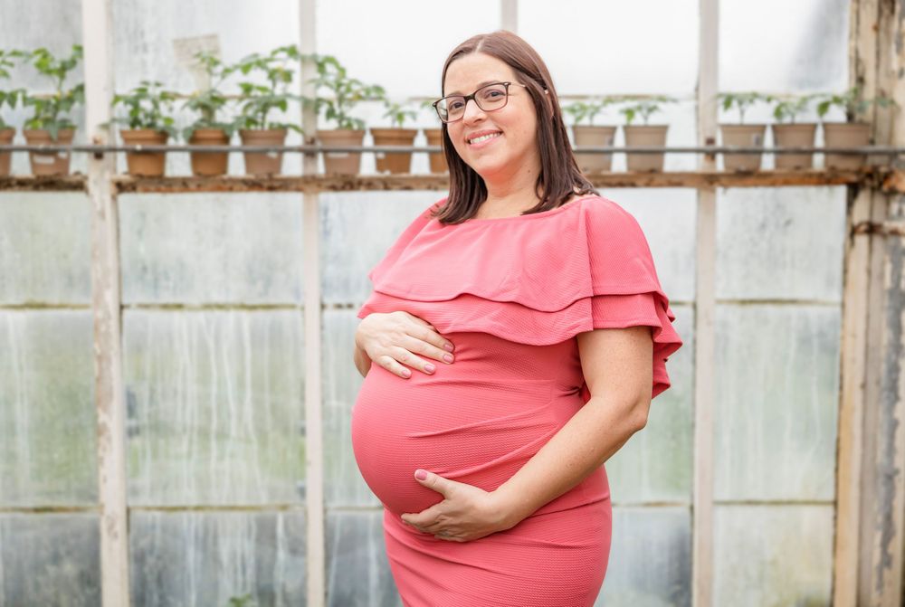 Expectant mother in coral ruffle dress smiles while standing in greenhouse.