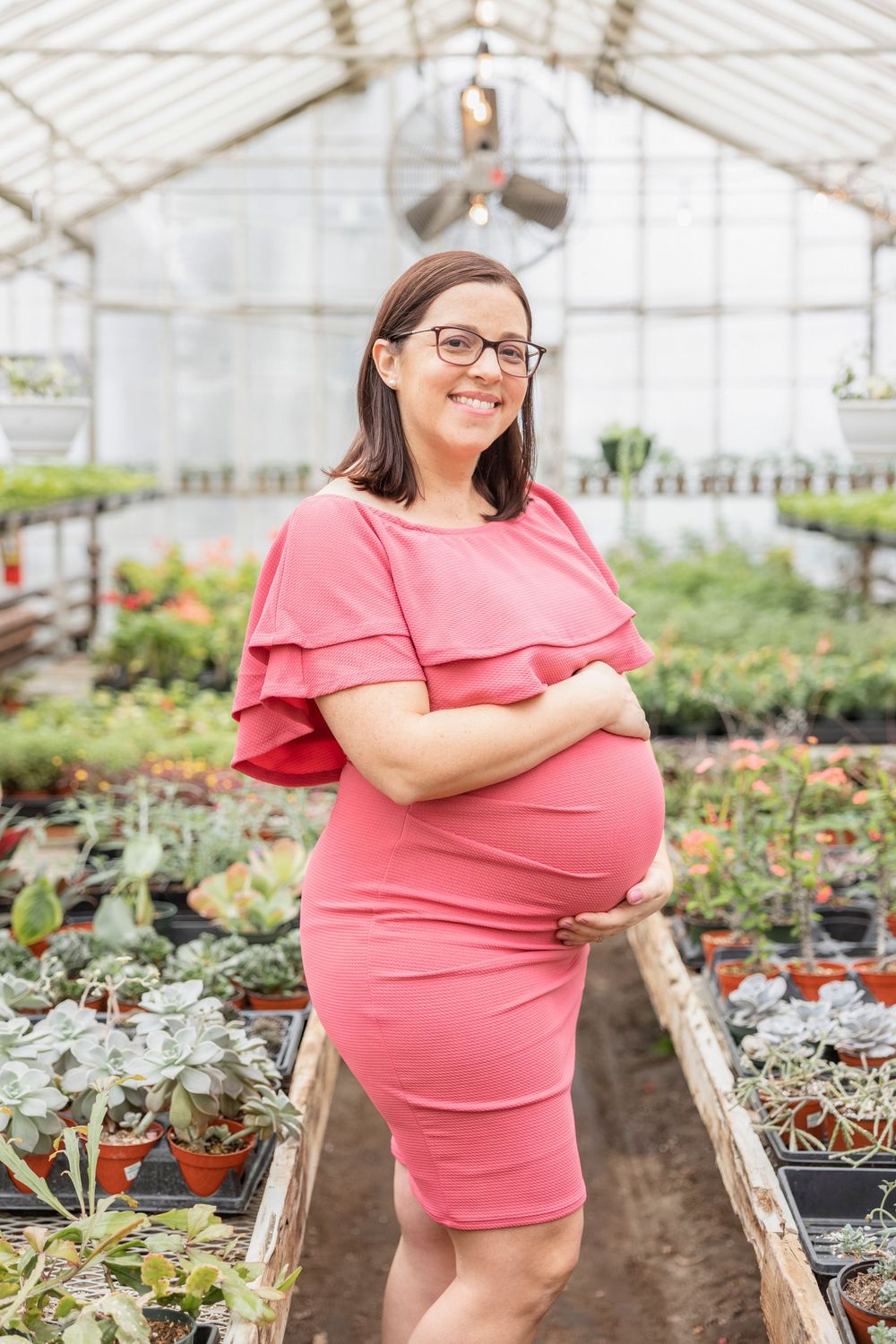 Pregnant woman in coral dress smiles while standing among plants in greenhouse setting.