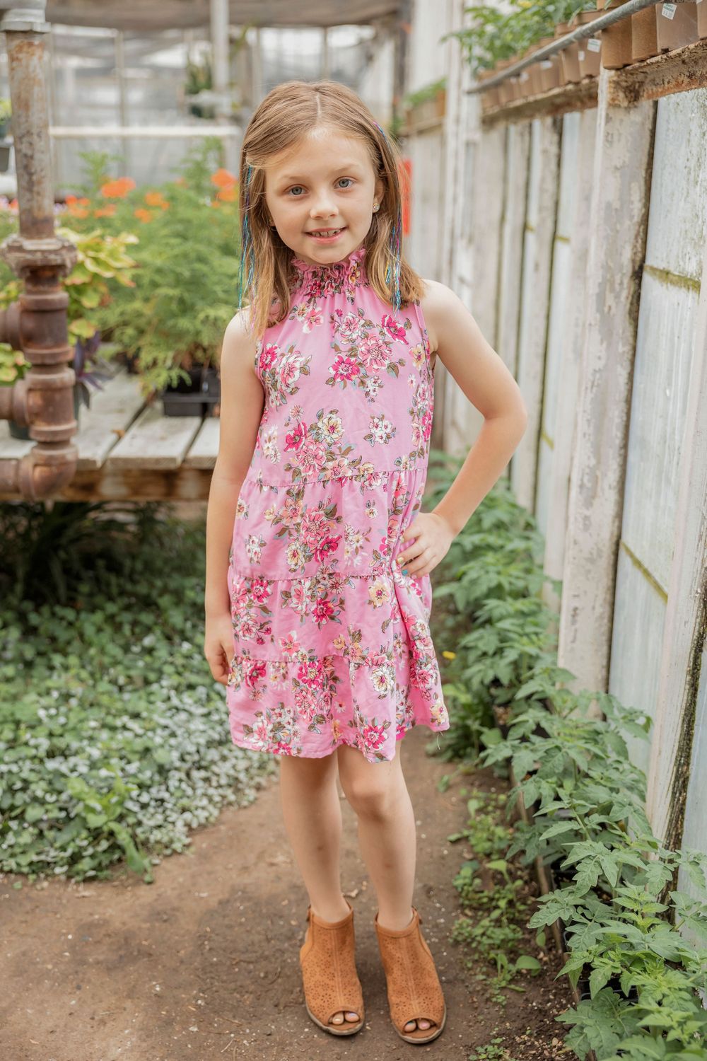 Young girl in pink floral dress poses confidently in greenhouse garden path.