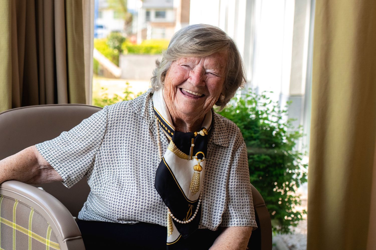 A joyful senior wearing a checkered shirt and necklace laughing while seated indoors.