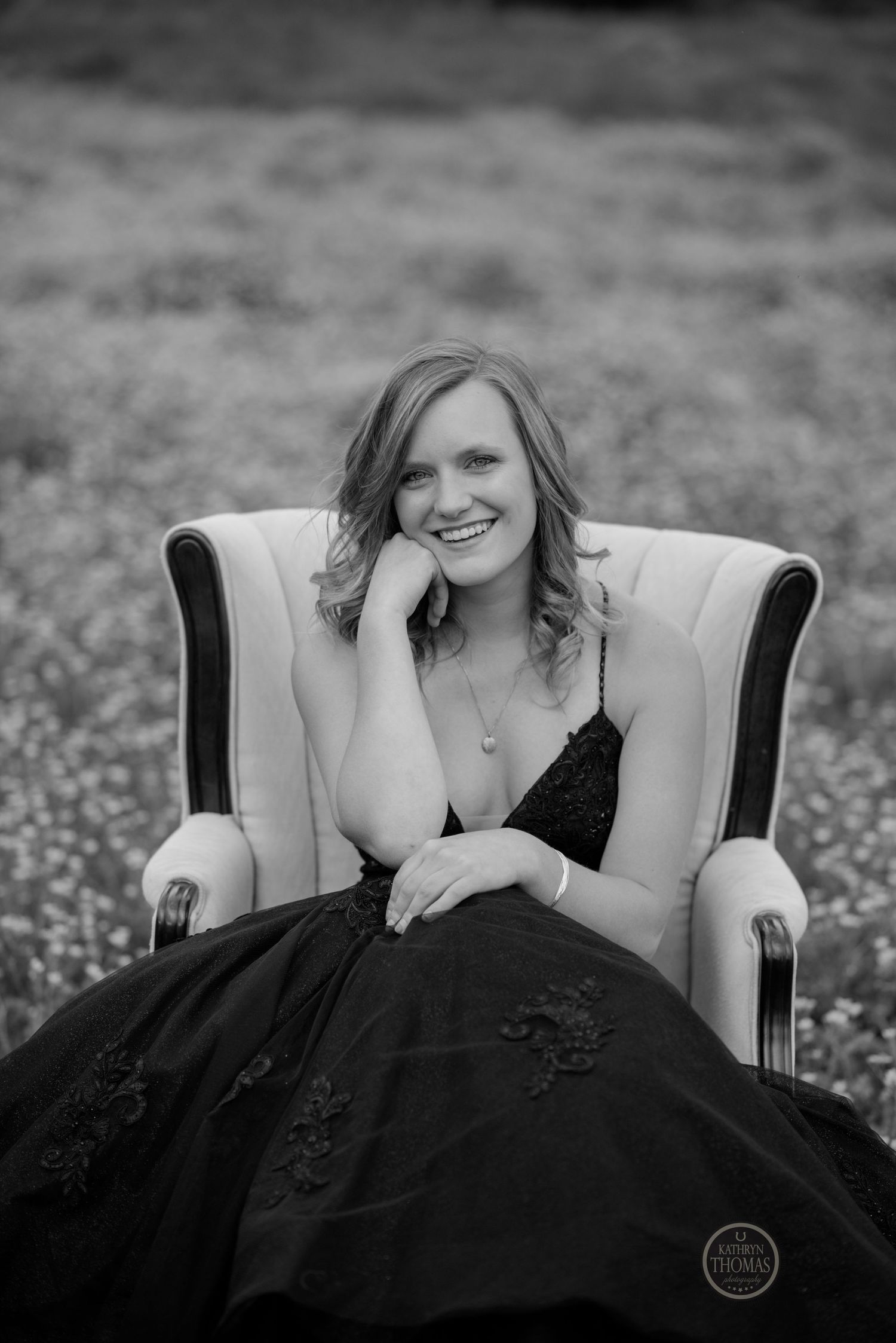A black and white portrait shows a figure in a black gown lounging in a beige armchair surrounded by wildflowers.