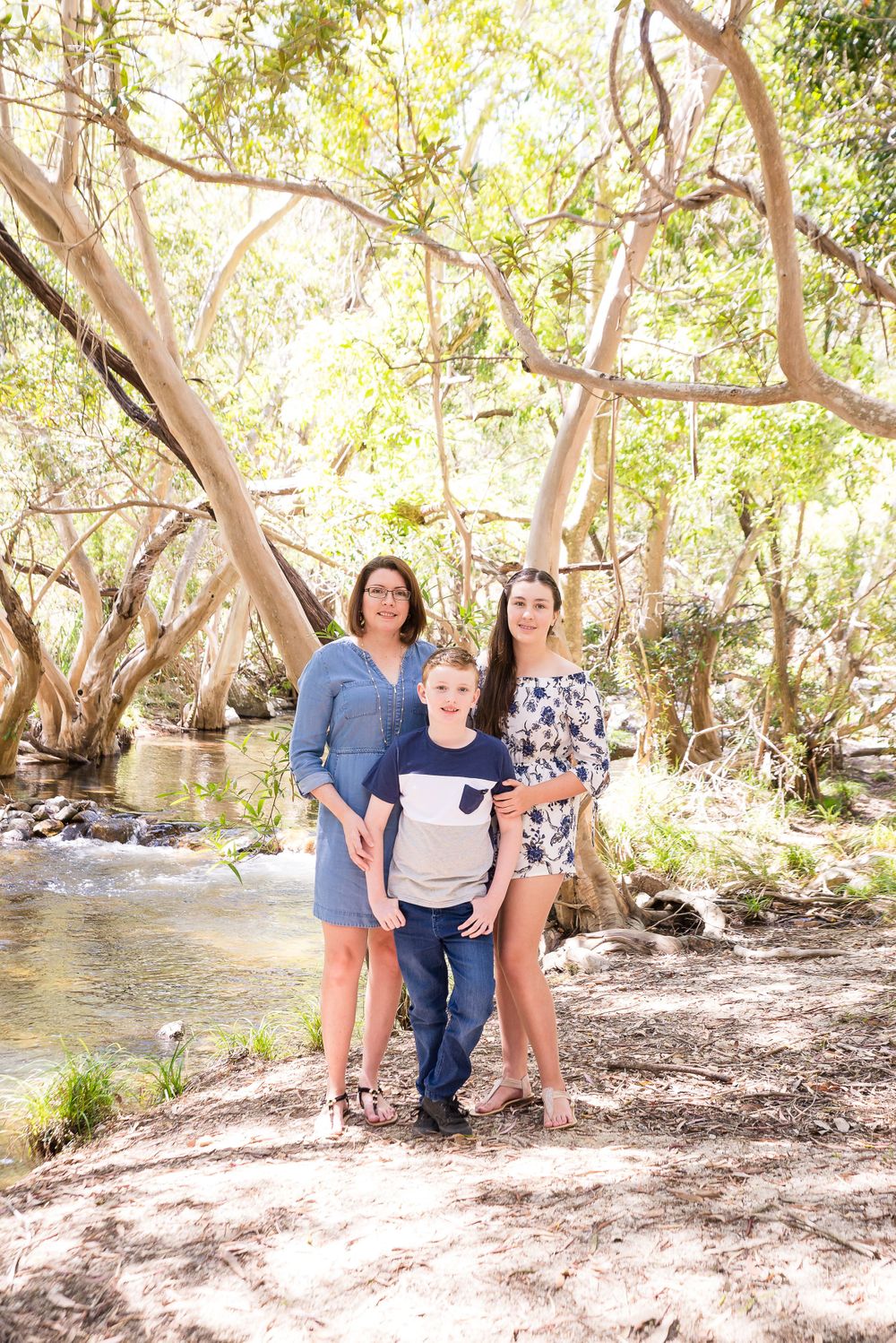 Family resting by creek bank with overhanging trees in sunlit natural setting.