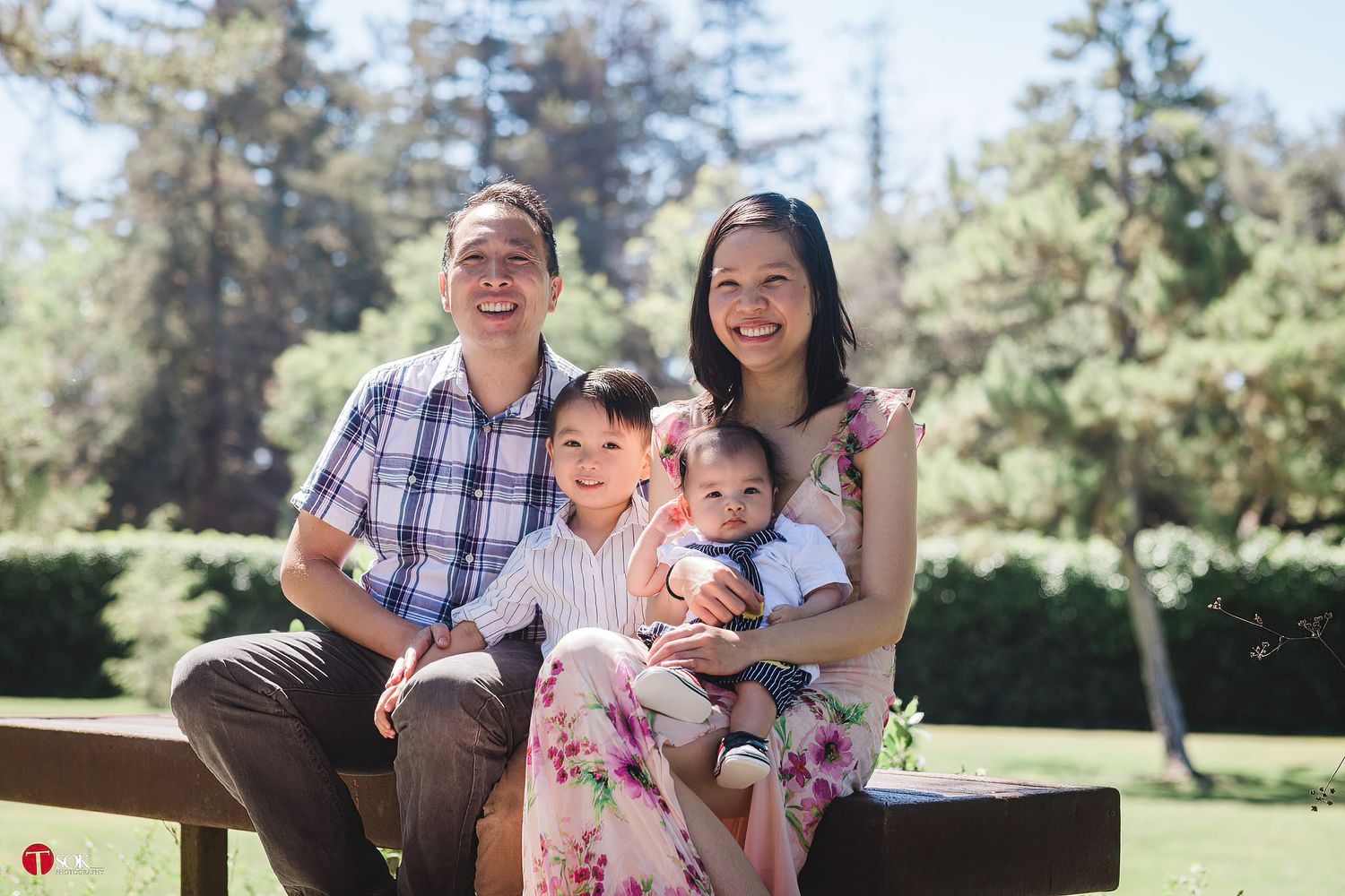 A family of four sits together on a wooden bench in a sunny garden setting.