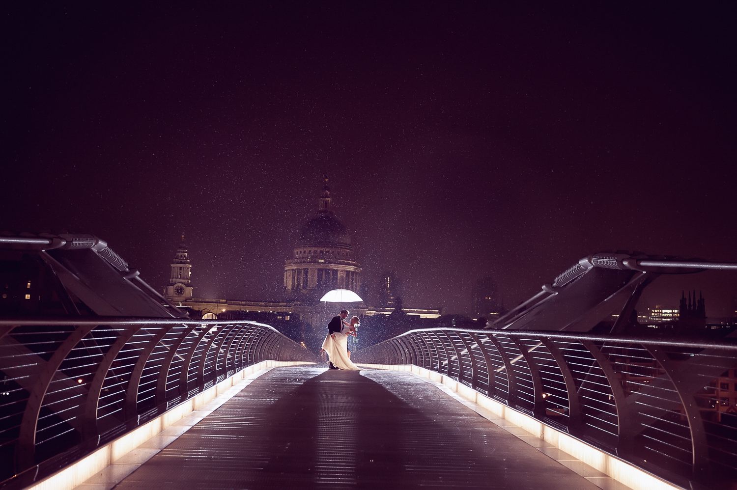 Night wedding photo on illuminated bridge with cathedral silhouette in purple haze.
