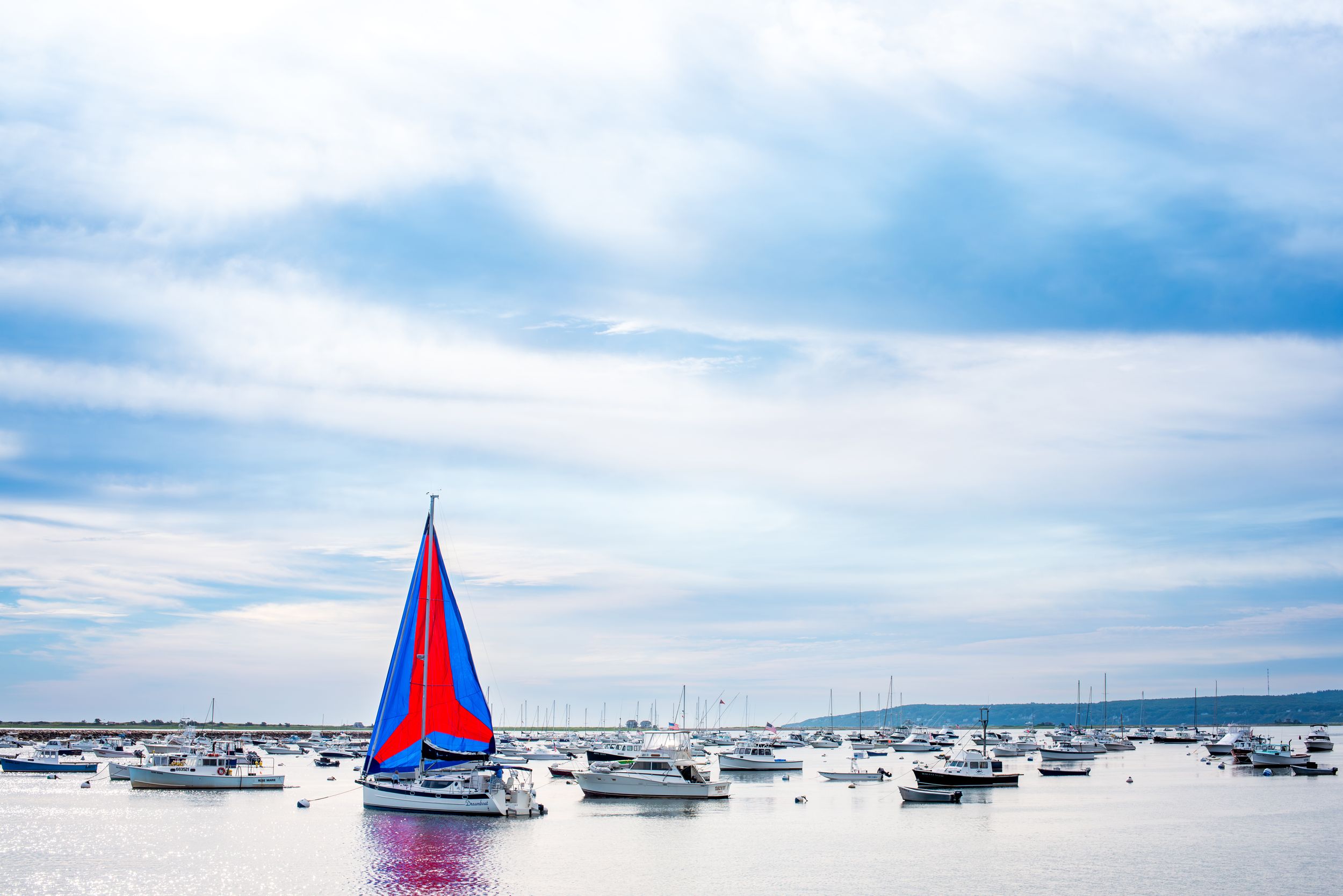 Colorful sailboat with red and blue sails anchored among other boats in a marina on a cloudy day.
