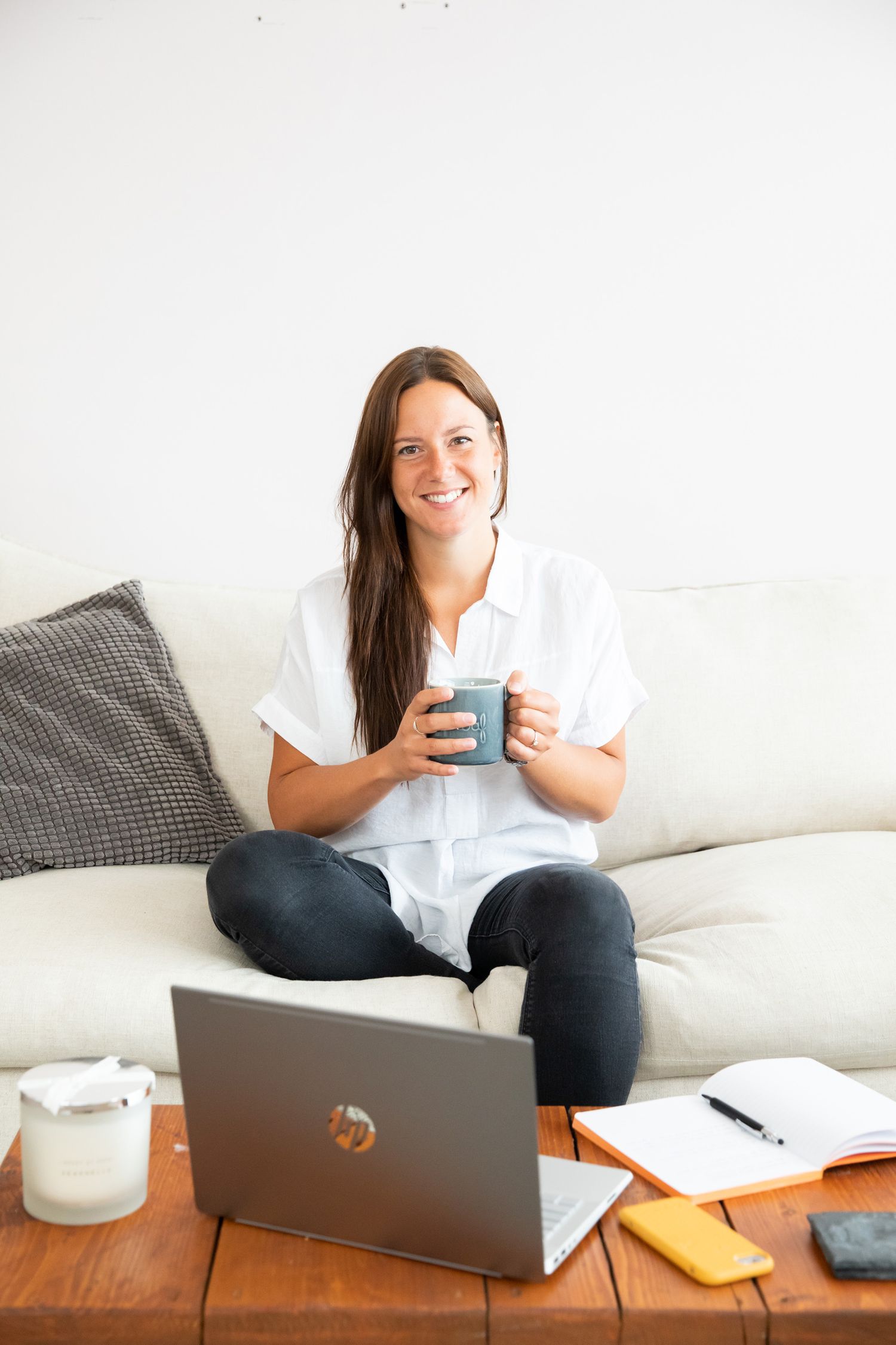 Individual relaxing on white couch with laptop and coffee mug in minimalist home office setting during business branding photoshoot.
