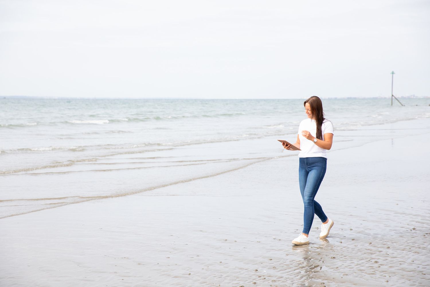Person walking along a serene beach shoreline while reading a book during business branding photoshoot.