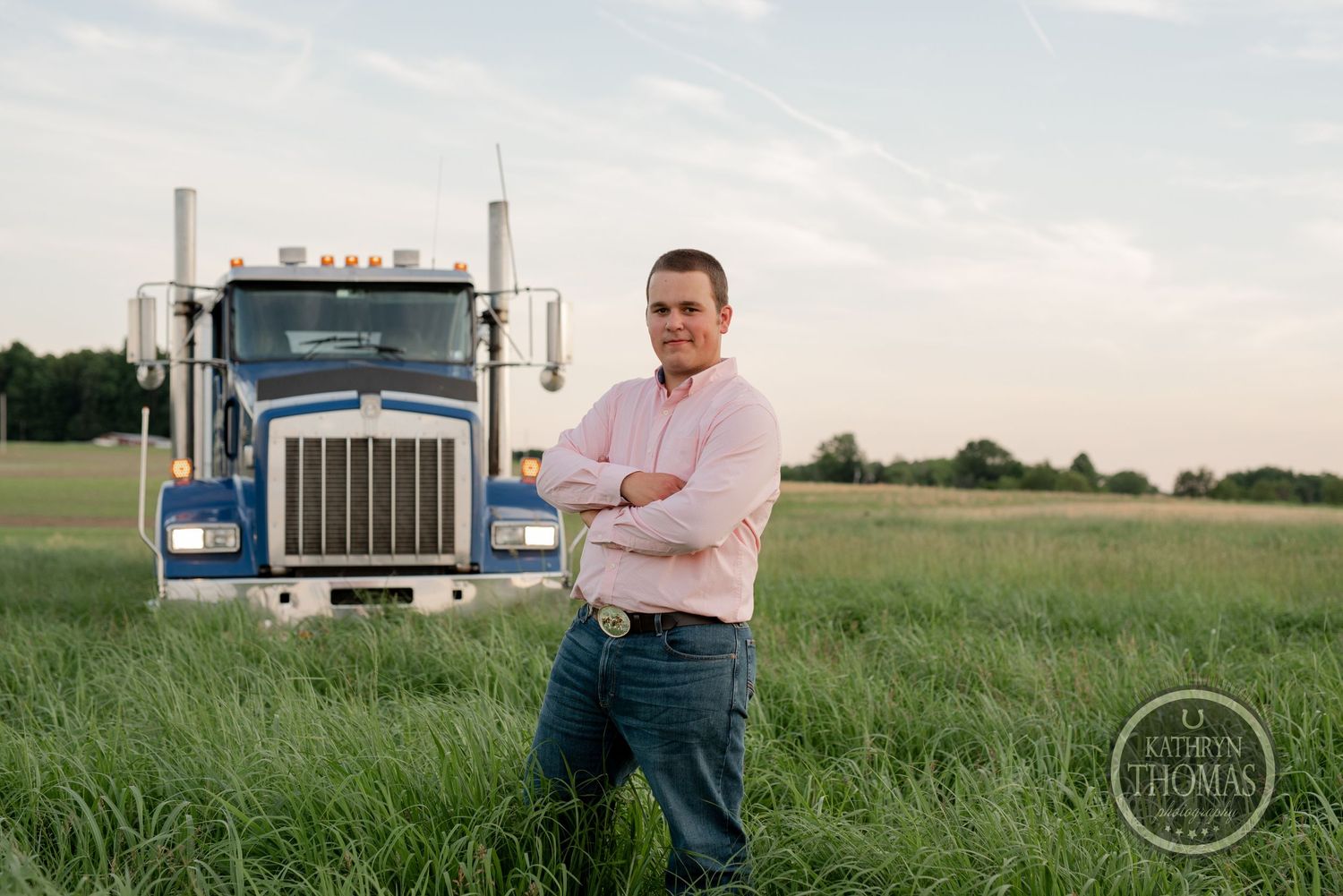 Driver stands with arms crossed beside blue semi truck in tall grass during golden hour.
