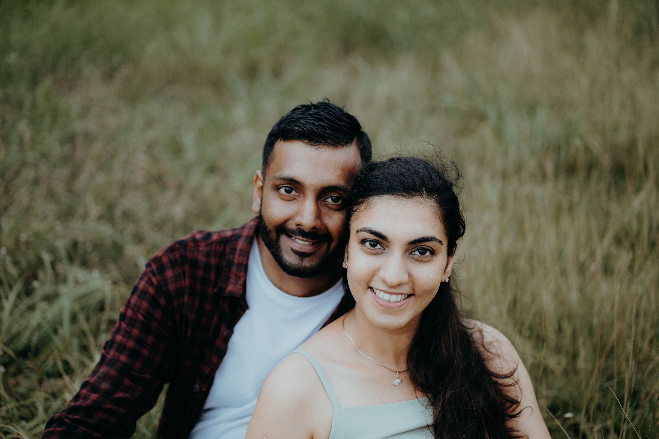 A couple in casual attire smiles while sitting together in a grassy field.
