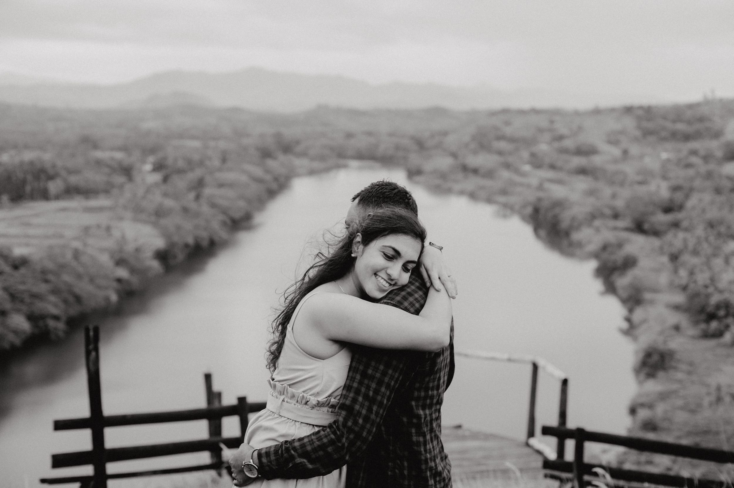 A black and white photo captures an embrace on a viewing platform overlooking a winding river.