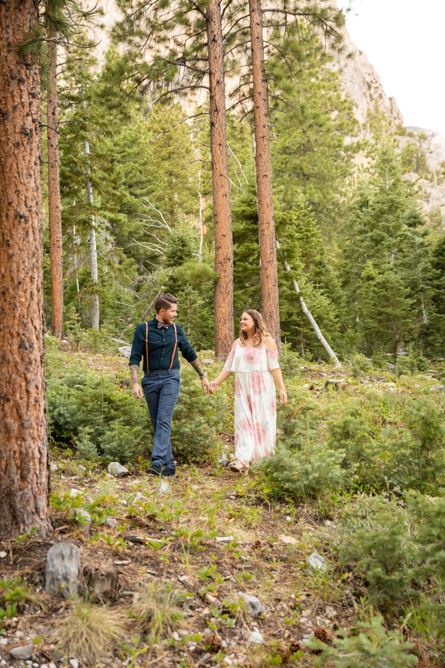 A couple walks hand in hand through a mountain forest trail surrounded by tall pine trees.