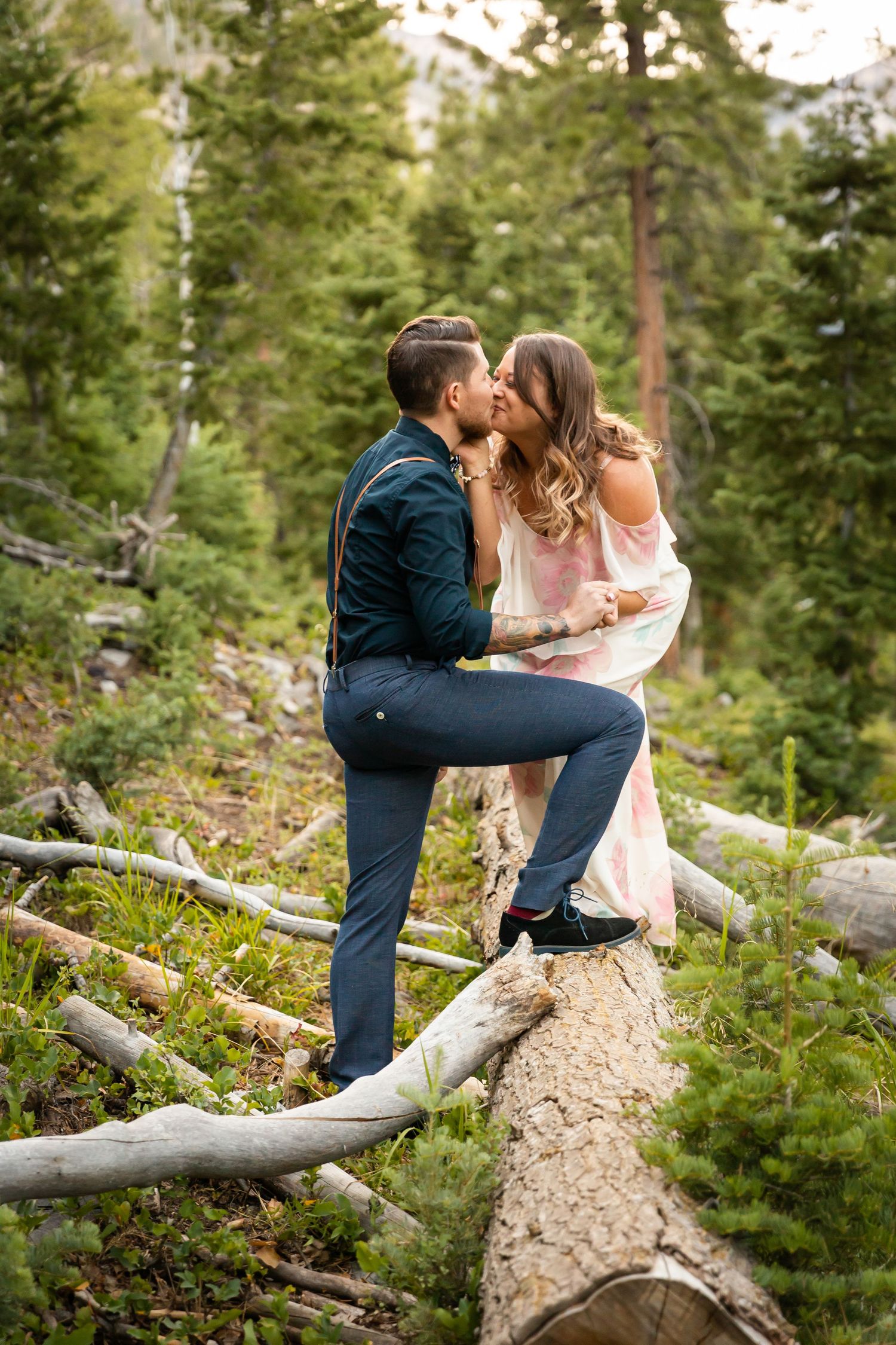 A couple shares an intimate moment while perched on a fallen tree trunk in a forest setting.