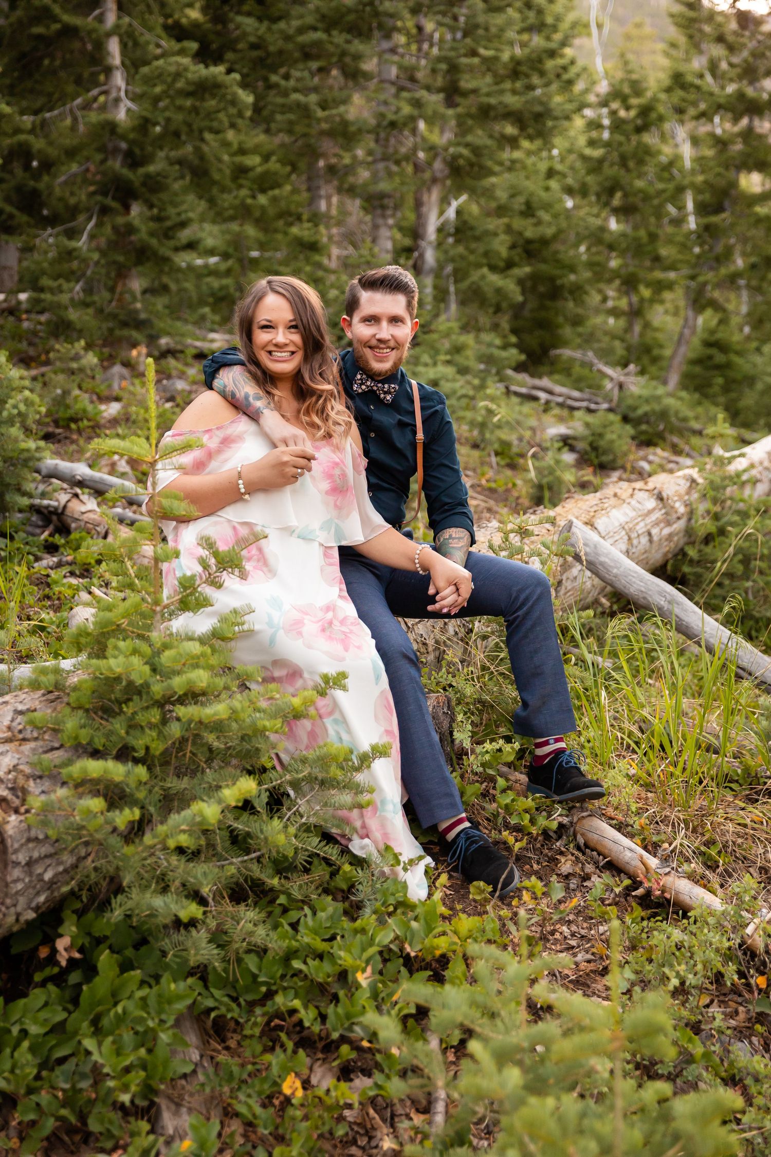 A romantic mountain photoshoot shows a couple sitting on a fallen log surrounded by evergreen trees.