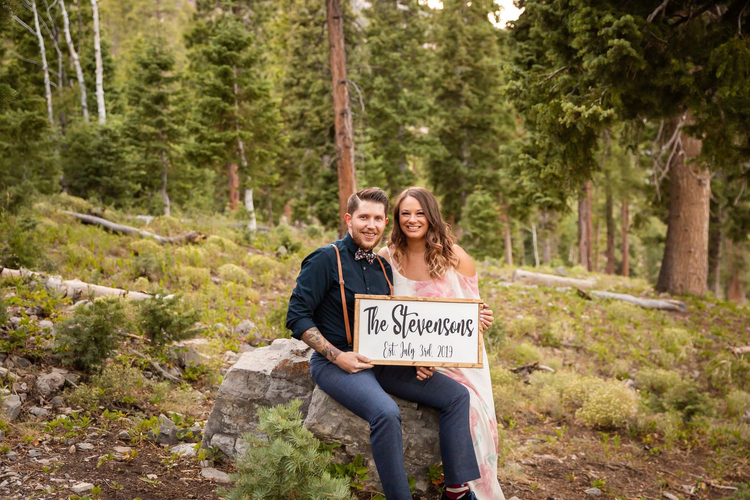 Couple sitting on log holding 'The Stevensons' sign in mountain forest landscape.
