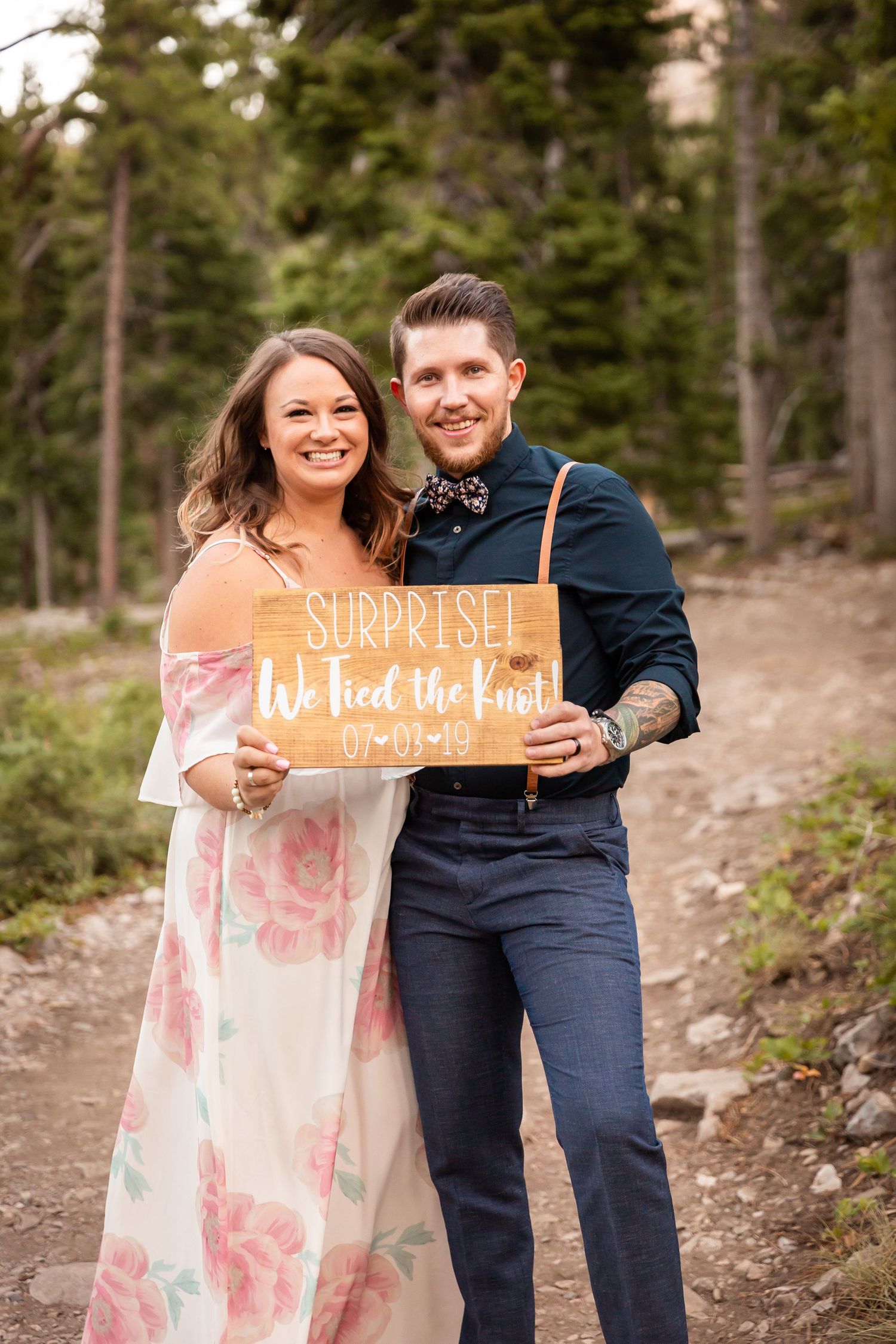 Couple holding sign reading 'Surprise We Tied the Knot' in mountain forest setting.