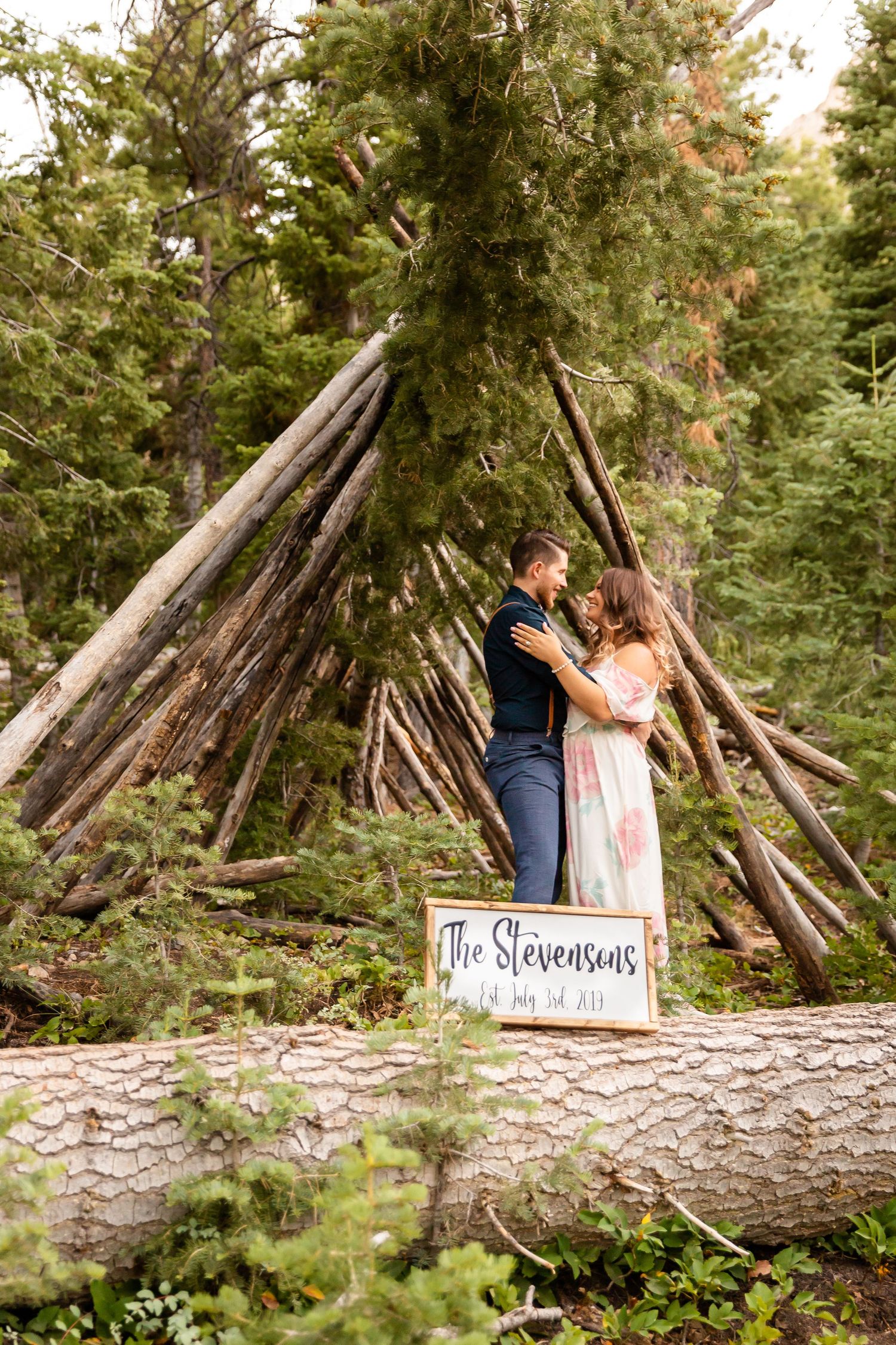 Couple poses with 'The Stevensons' sign in front of wooden teepee structure in forest.