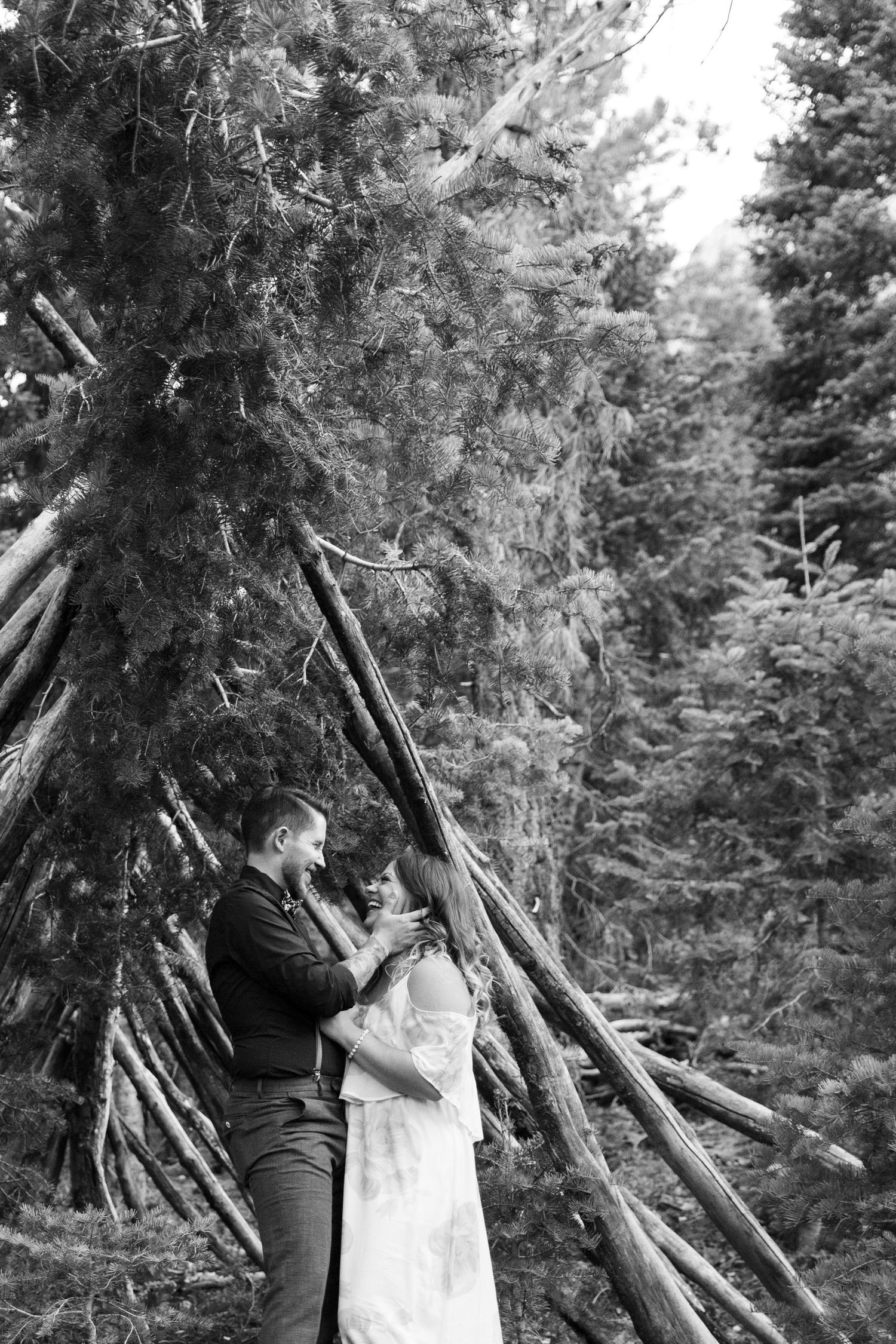 Black and white portrait of couple in romantic pose against forest teepee structure.
