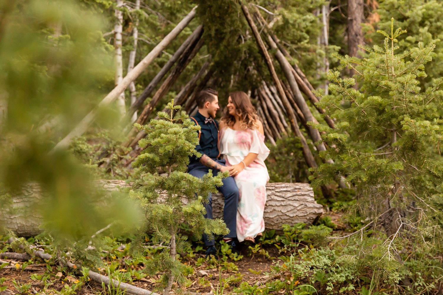 Happy couple posing together near wooden teepee structure surrounded by pine trees.
