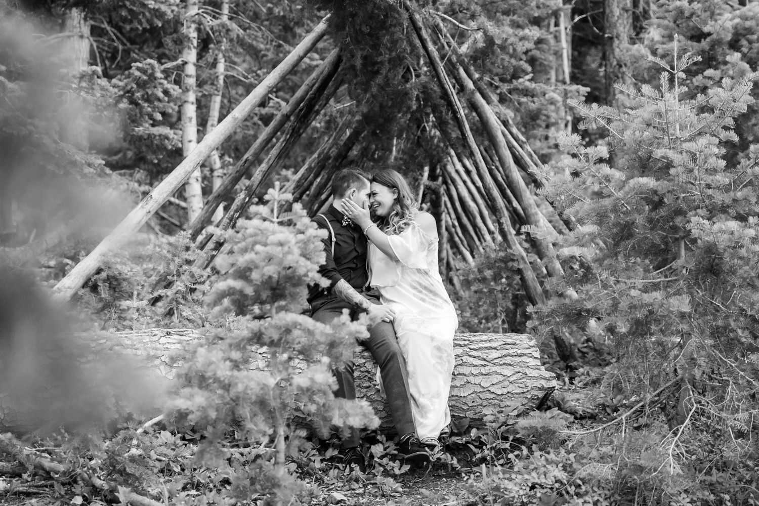 Black and white photo of couple sitting by rustic wooden teepee structure in forest setting.