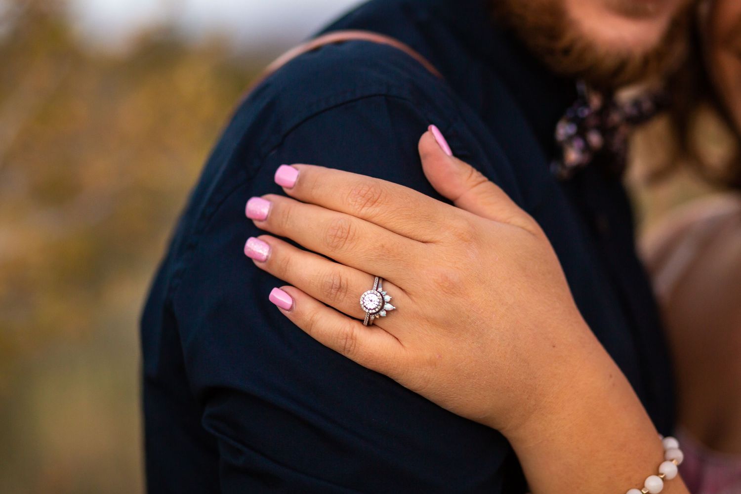 Close-up of a hand with pink nail polish displaying a sparkling engagement ring against a dark sleeve.