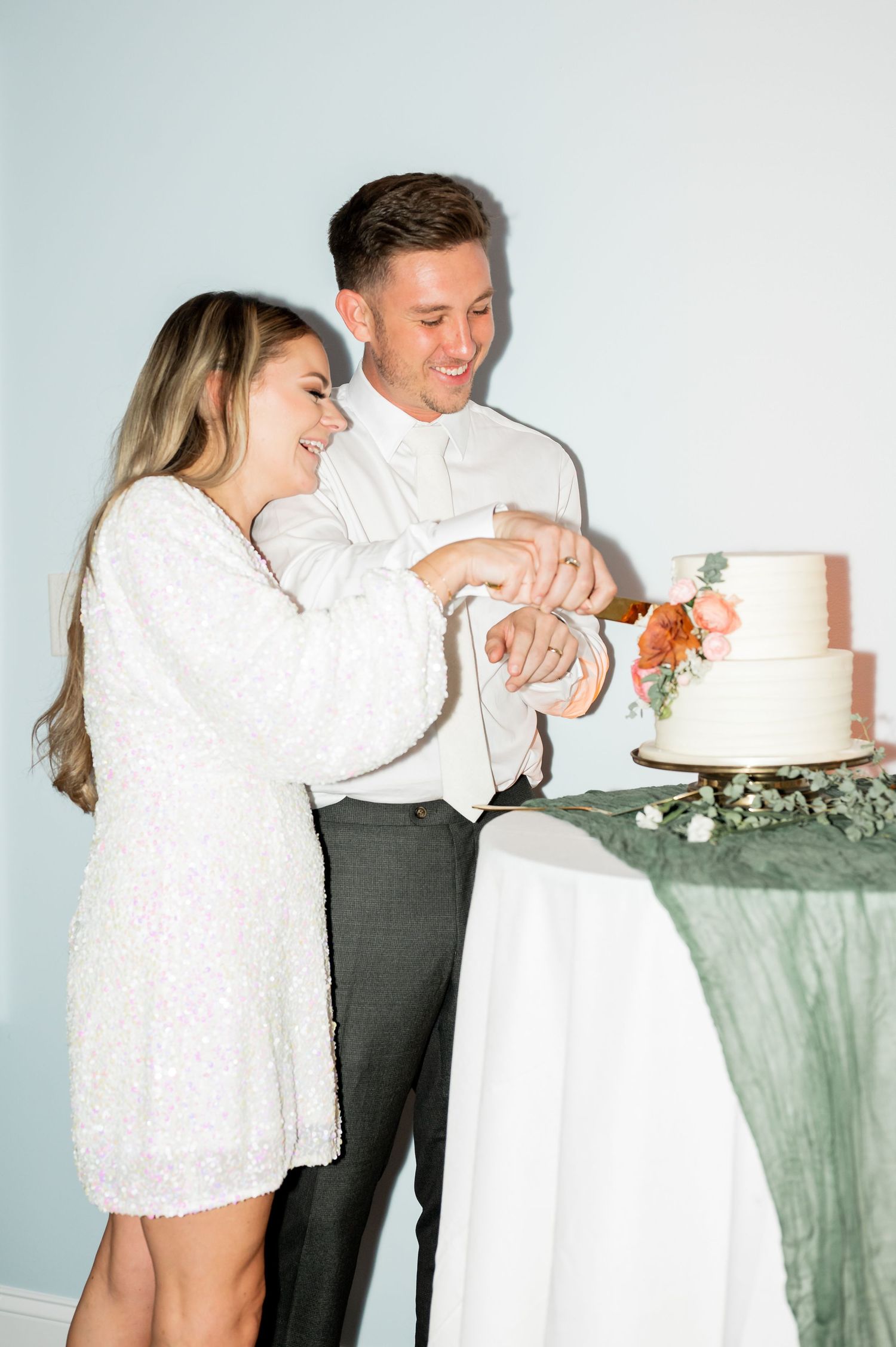 Couple in white attire sharing laughter while cutting their wedding cake together.