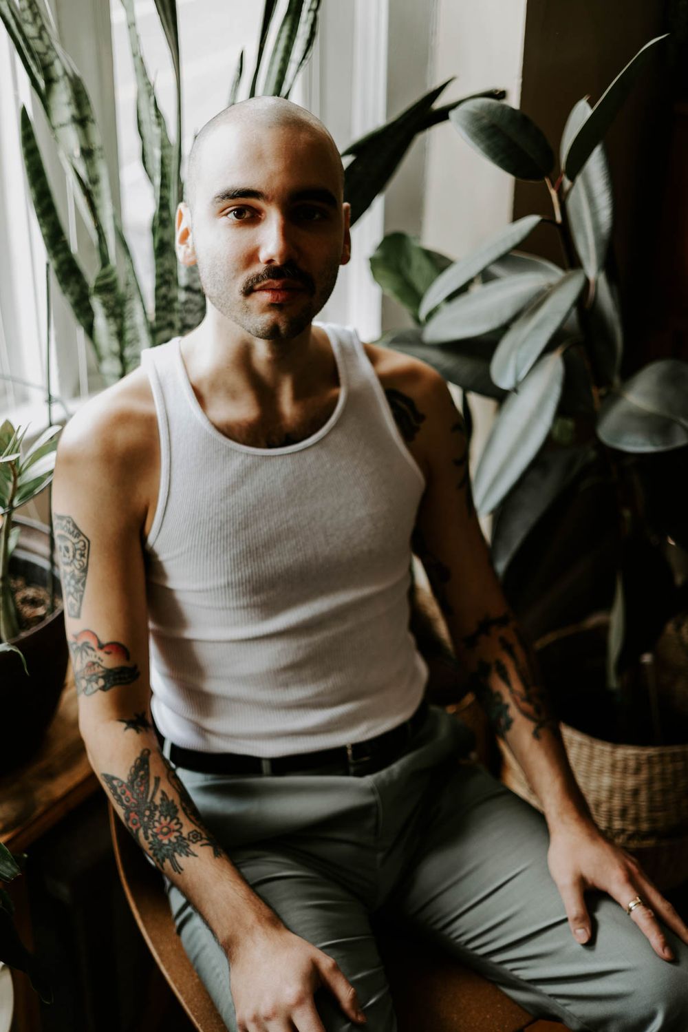 Person in white tank top and gray pants seated near indoor plants by window.