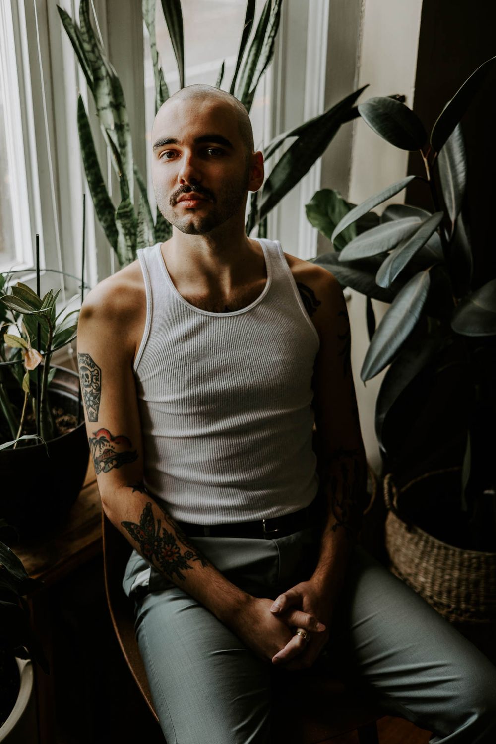 Moody portrait of person in tank top with tattoos sitting by houseplants near window.