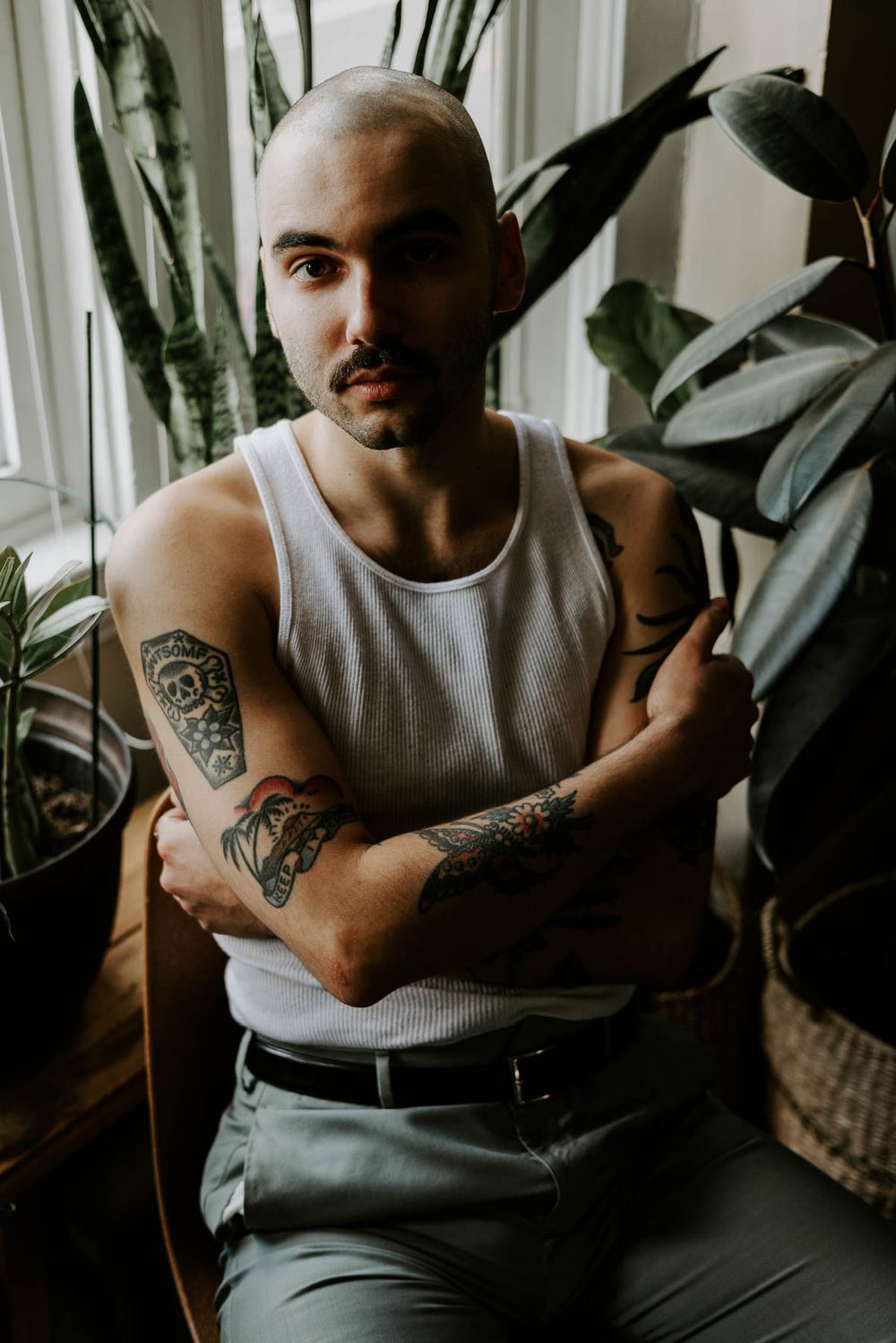 Person with tattoos in white tank top poses near window with indoor plants.