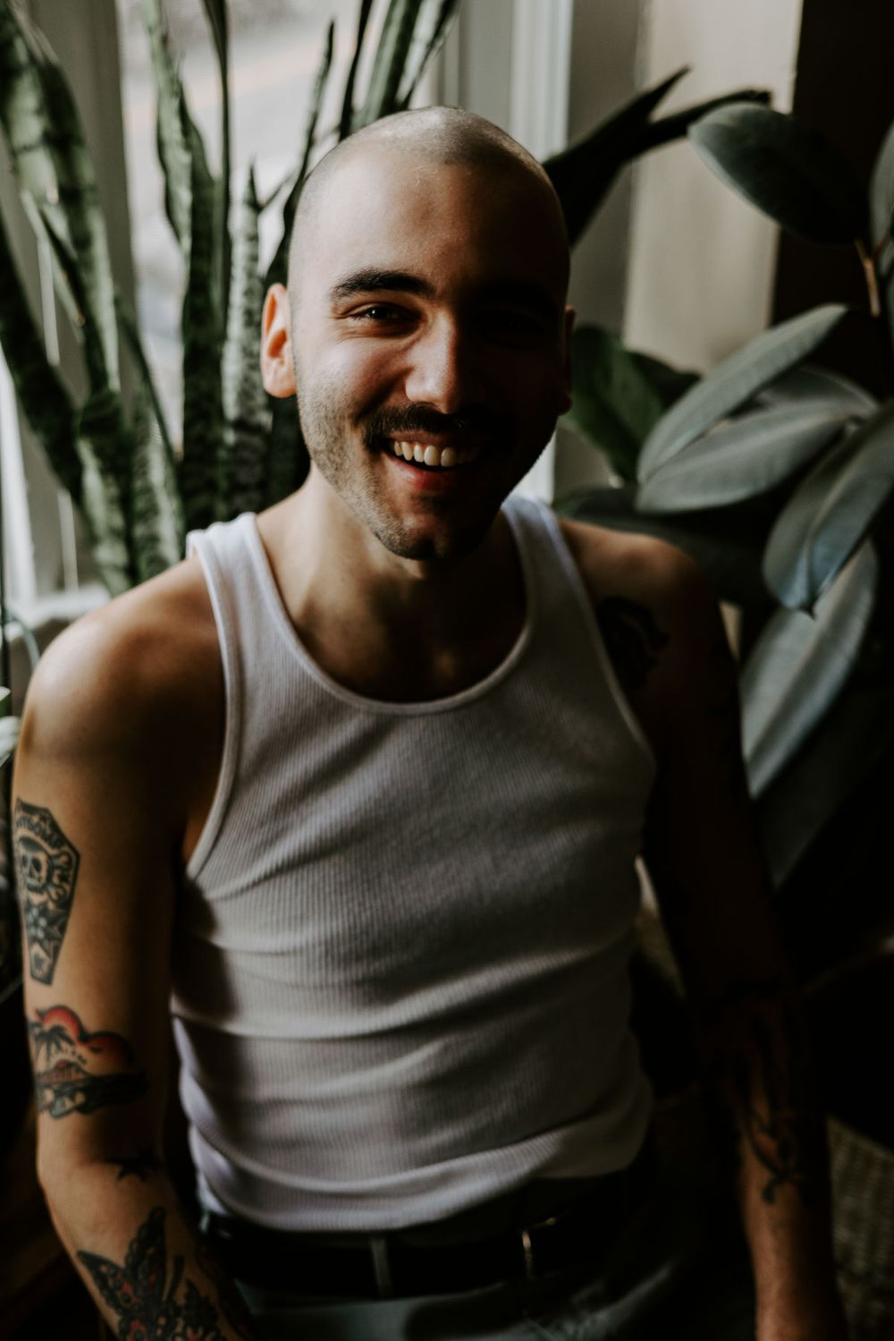 Person in white tank top smiling while sitting near green plants in natural lighting.