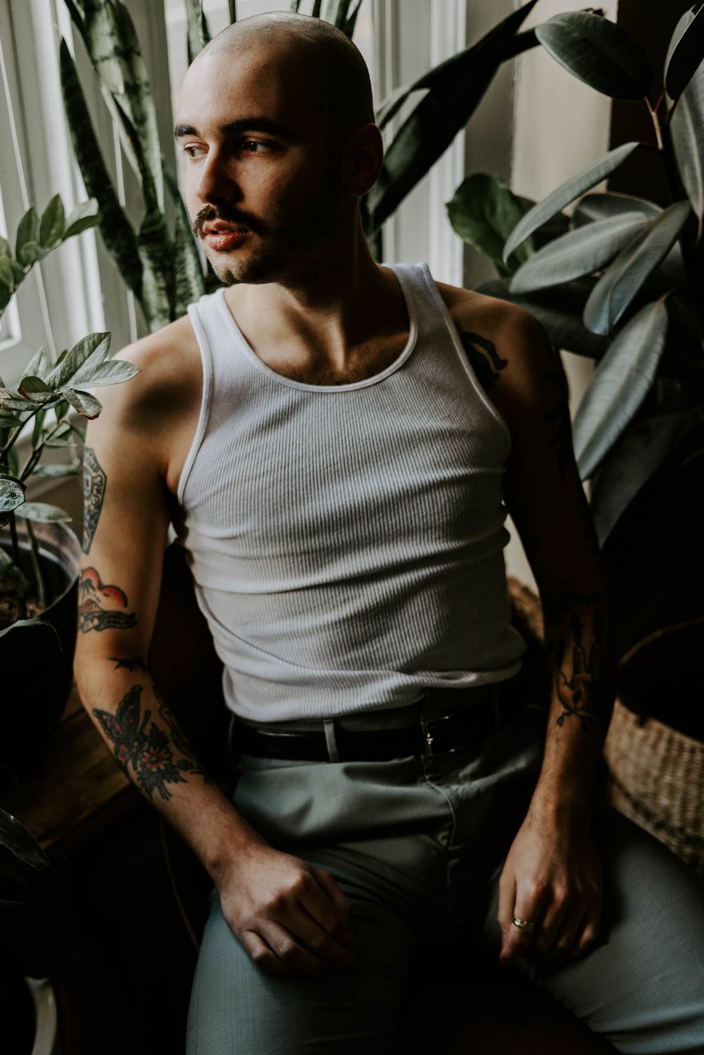Person wearing white tank top and dark pants seated by indoor plants.