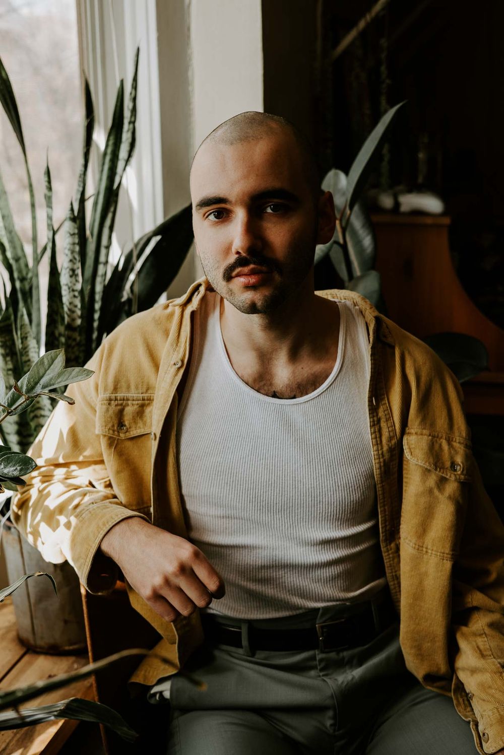 Figure in tan jacket and white tank top poses near window with houseplants.