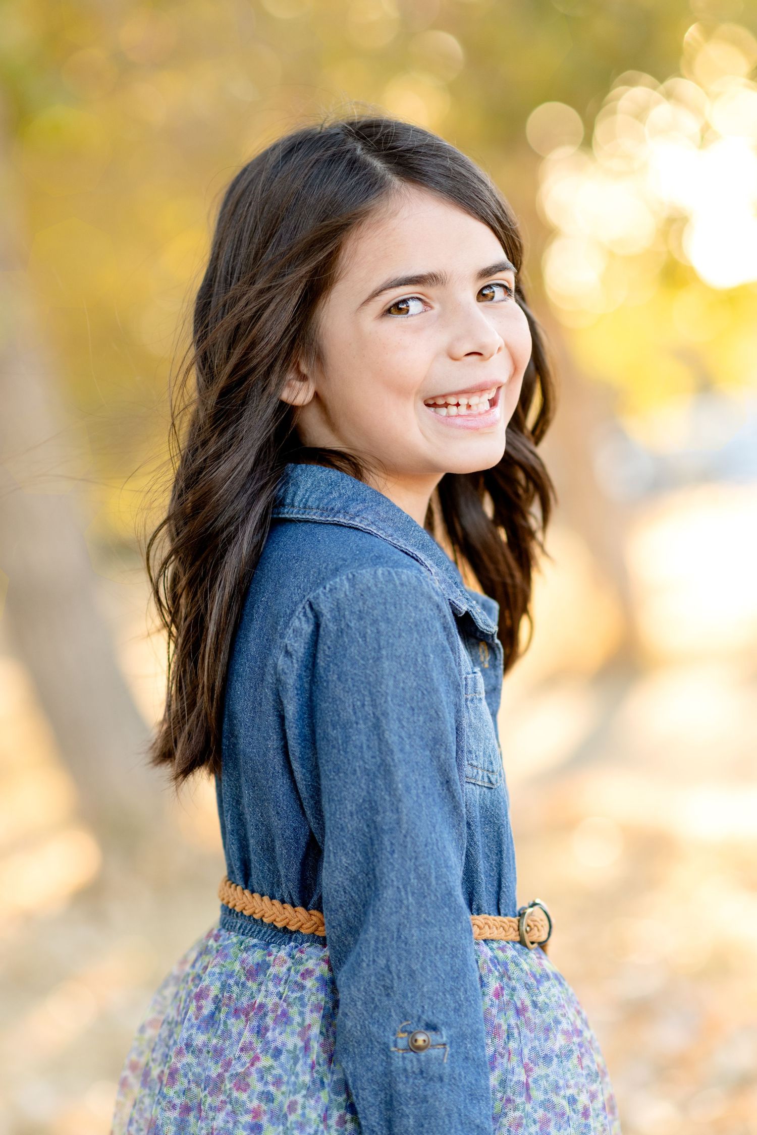 Person in denim jacket and floral dress poses with warm autumn bokeh background.
